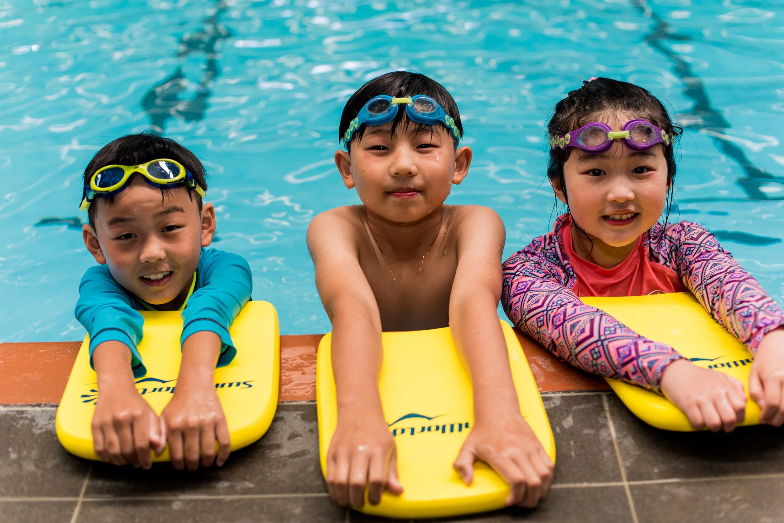 Three children in swimming goggles and swimwear, holding yellow flotation devices, at the edge of a swimming pool.