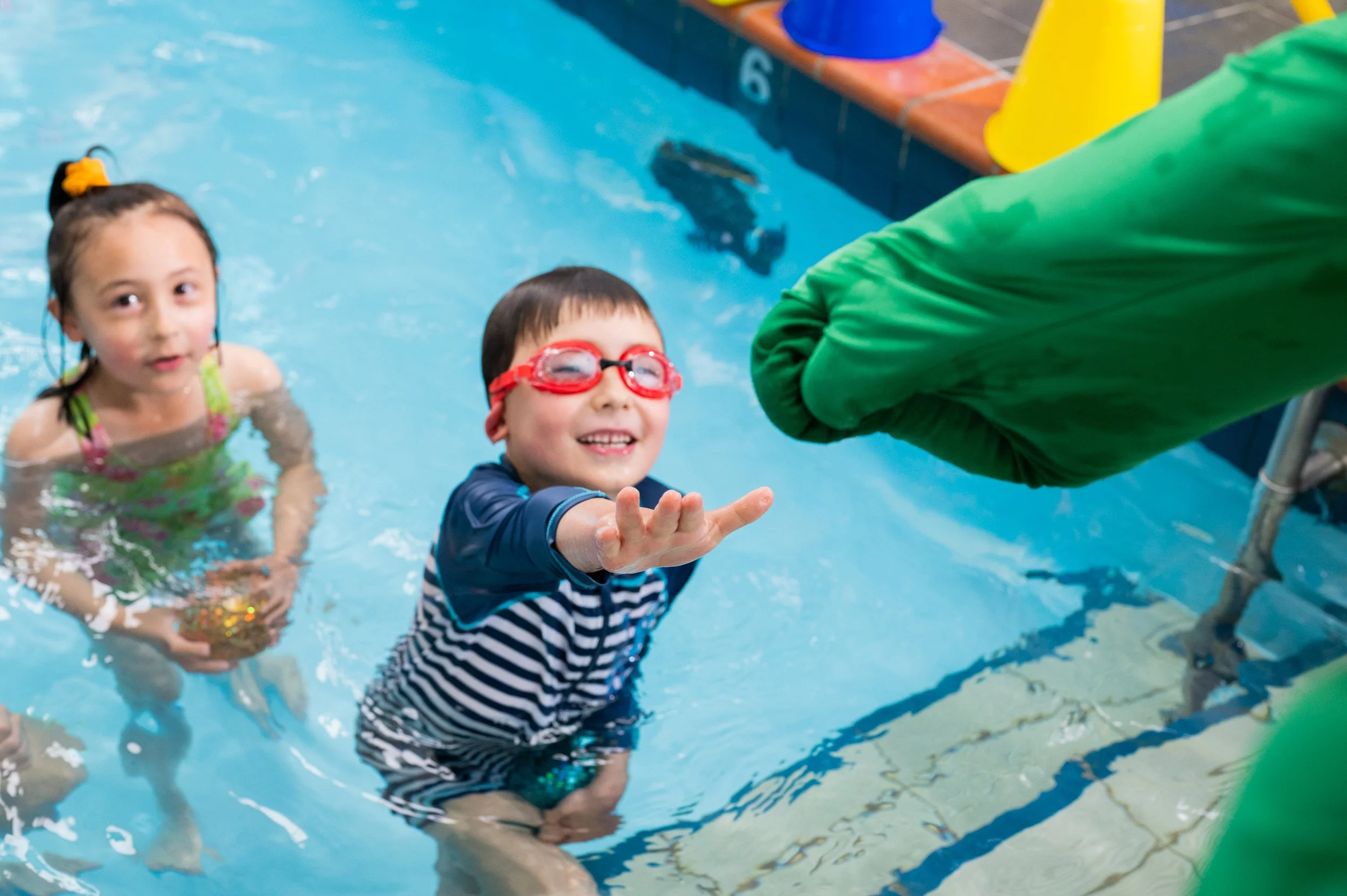 Two young children in a swimming pool, a girl and a boy, with the boy reaching out to a person in a green wetsuit. The boy is smiling and wearing red goggles, and the girl is holding a gold ball.