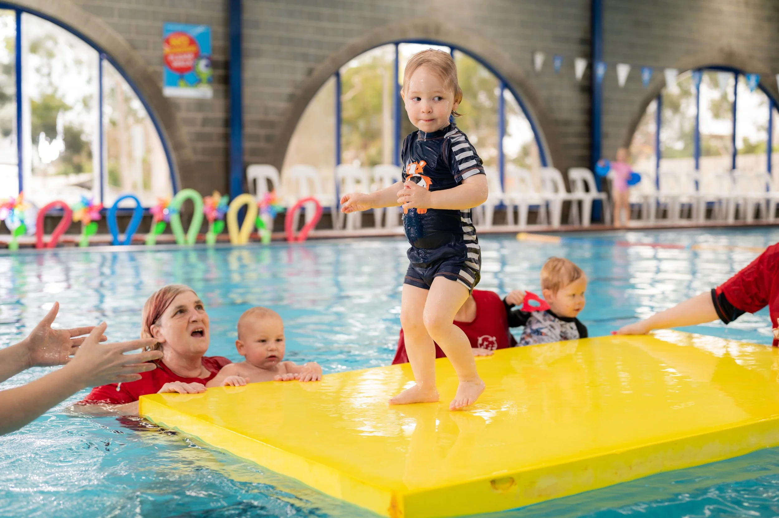 A young child standing on a yellow floating mat in an indoor swimming pool, with an adult and other children in the water nearby. The background includes colorful pool toys and chairs.