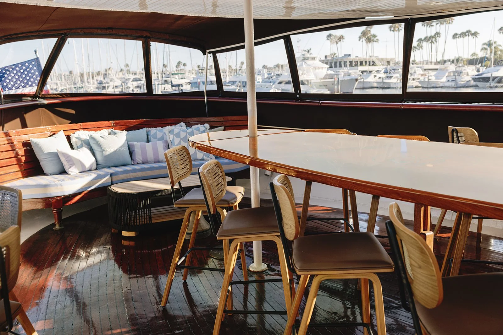 Outdoor seating area on a boat with a view of the marina and boats, American flag in the background, wooden deck, chairs, and tables.