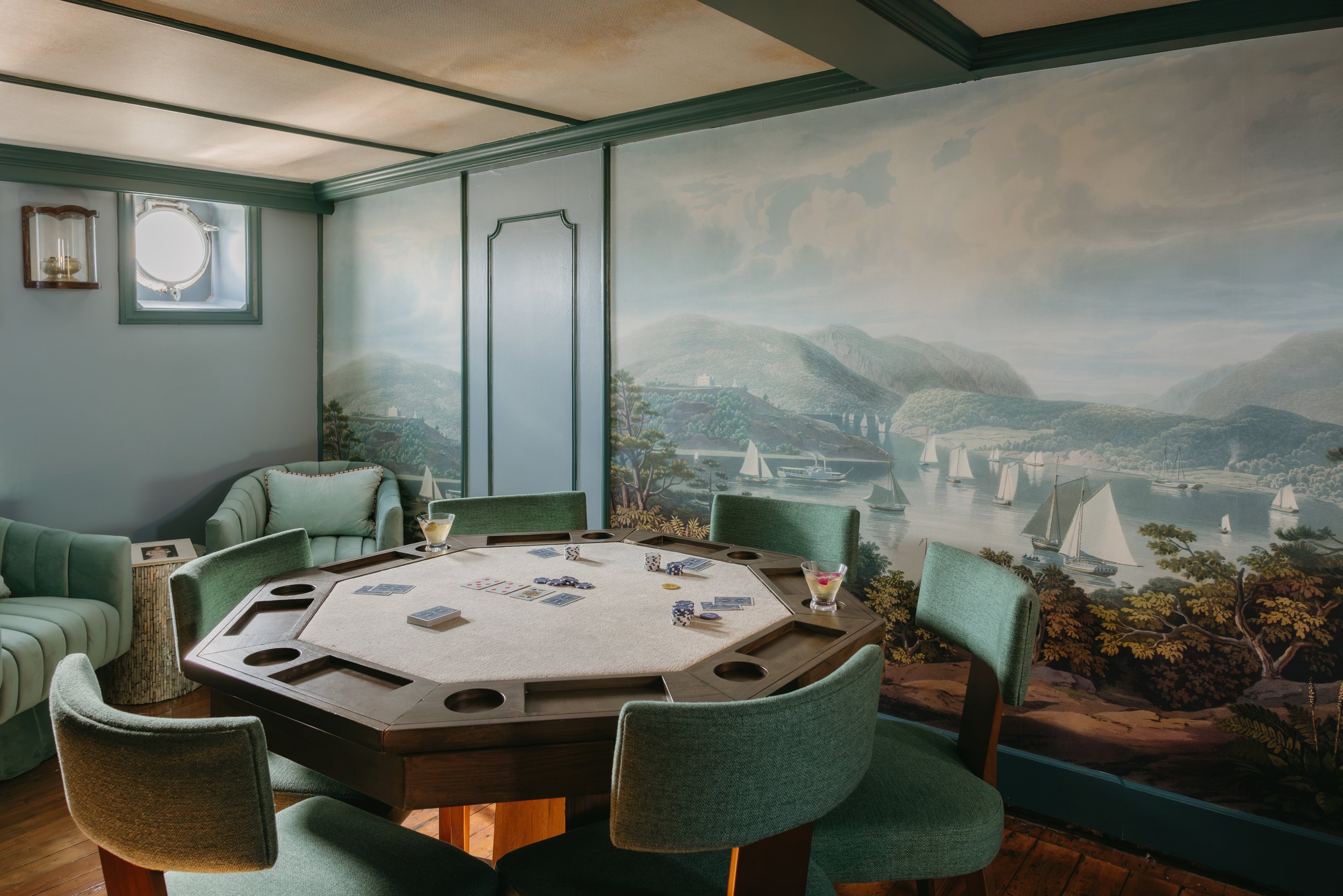 Room with a painted wall mural of a lake with sailboats, a round poker table with green chairs, a white armchair, a potted plant, and wall-mounted lamps.