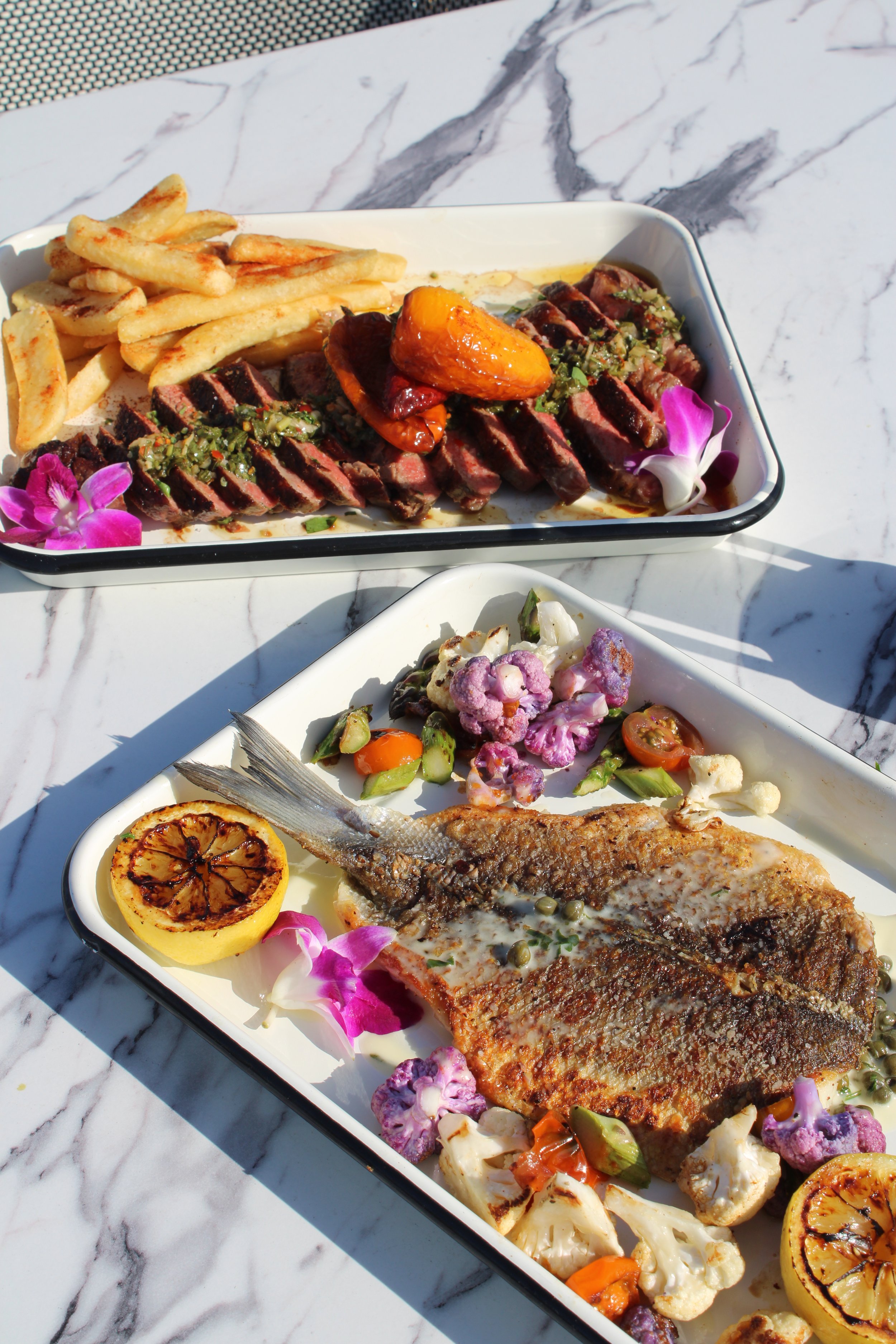 Two plates of grilled seafood with vegetables, lemon, and garnishes on a marble table, with one plate containing a grilled fish and the other containing sliced beef steak with sides.