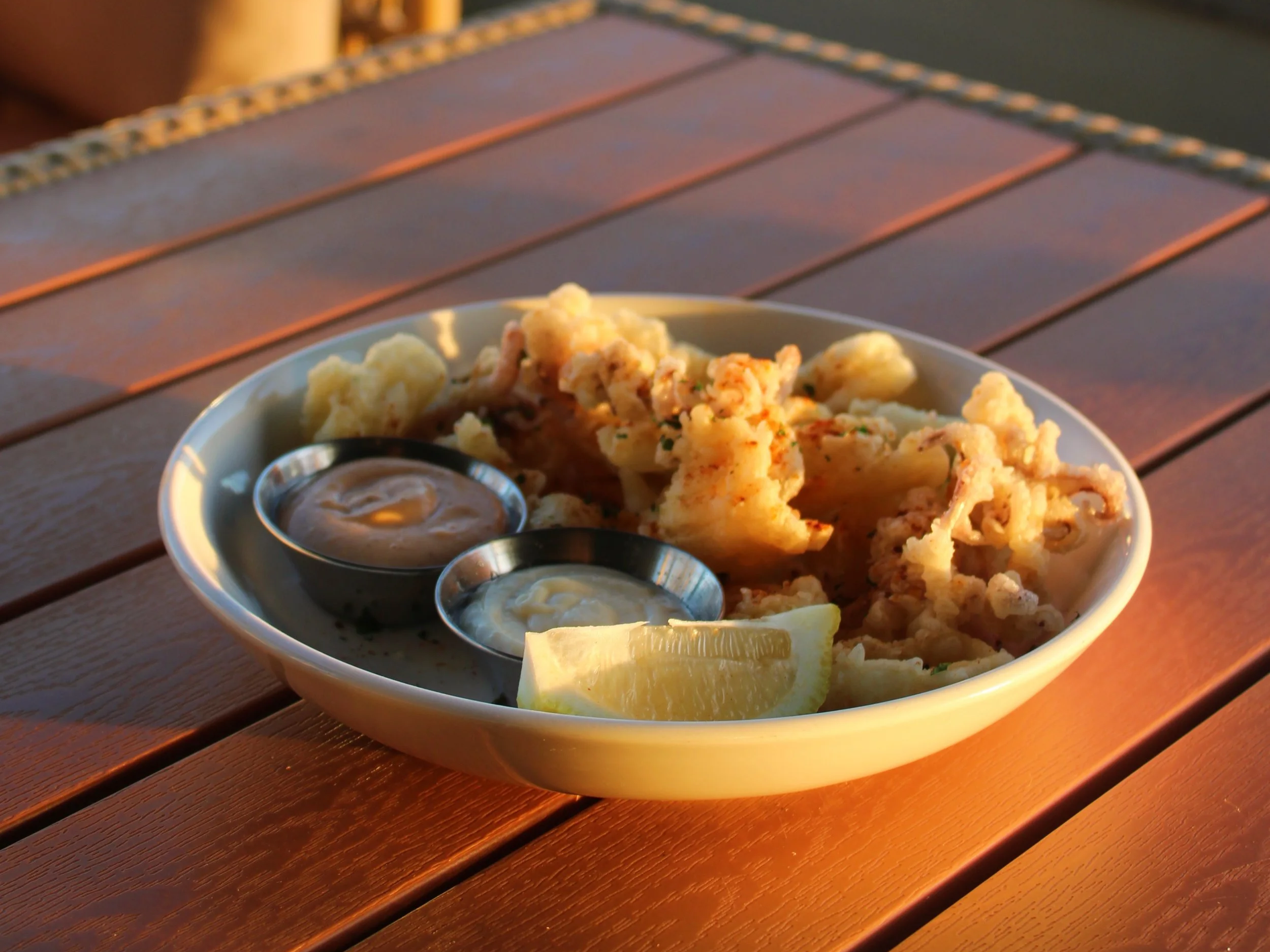 A white bowl of fried calamari with two dipping sauces and a lemon wedge on a wooden table.