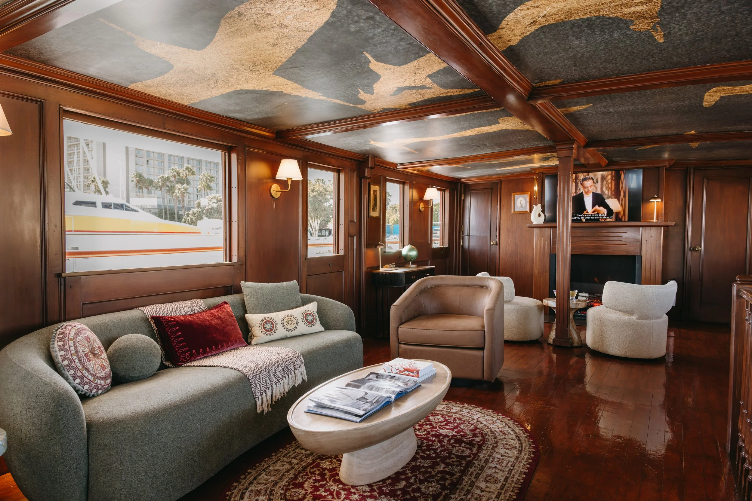 Interior of a wooden-paneled lounge room with a mural of fish on the ceiling, multiple windows, a fireplace, and a sitting area with sofas, armchairs, and a small table.