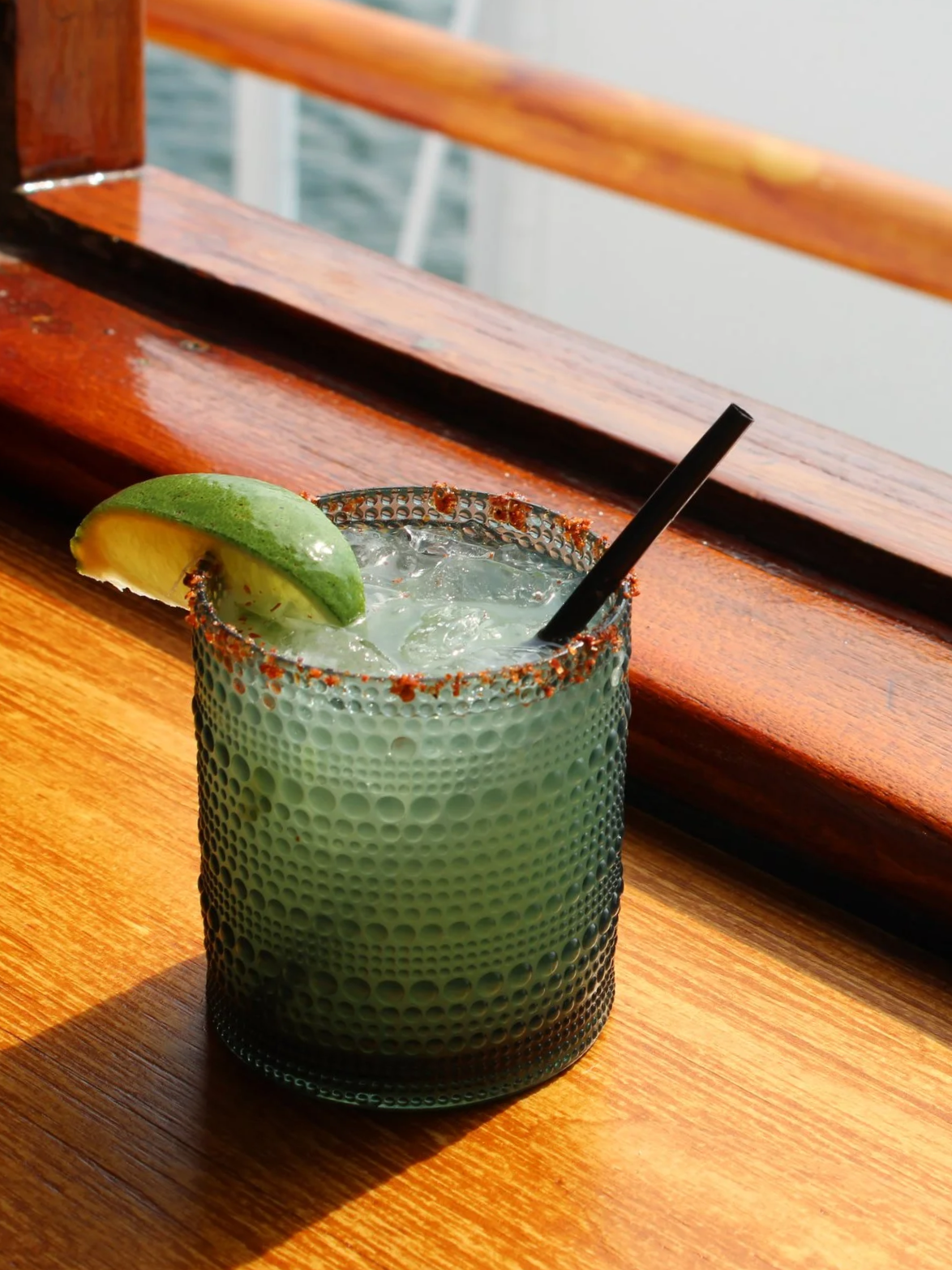 A green cocktail drink with a lime wedge and chili powder rim on a wooden table near the water.