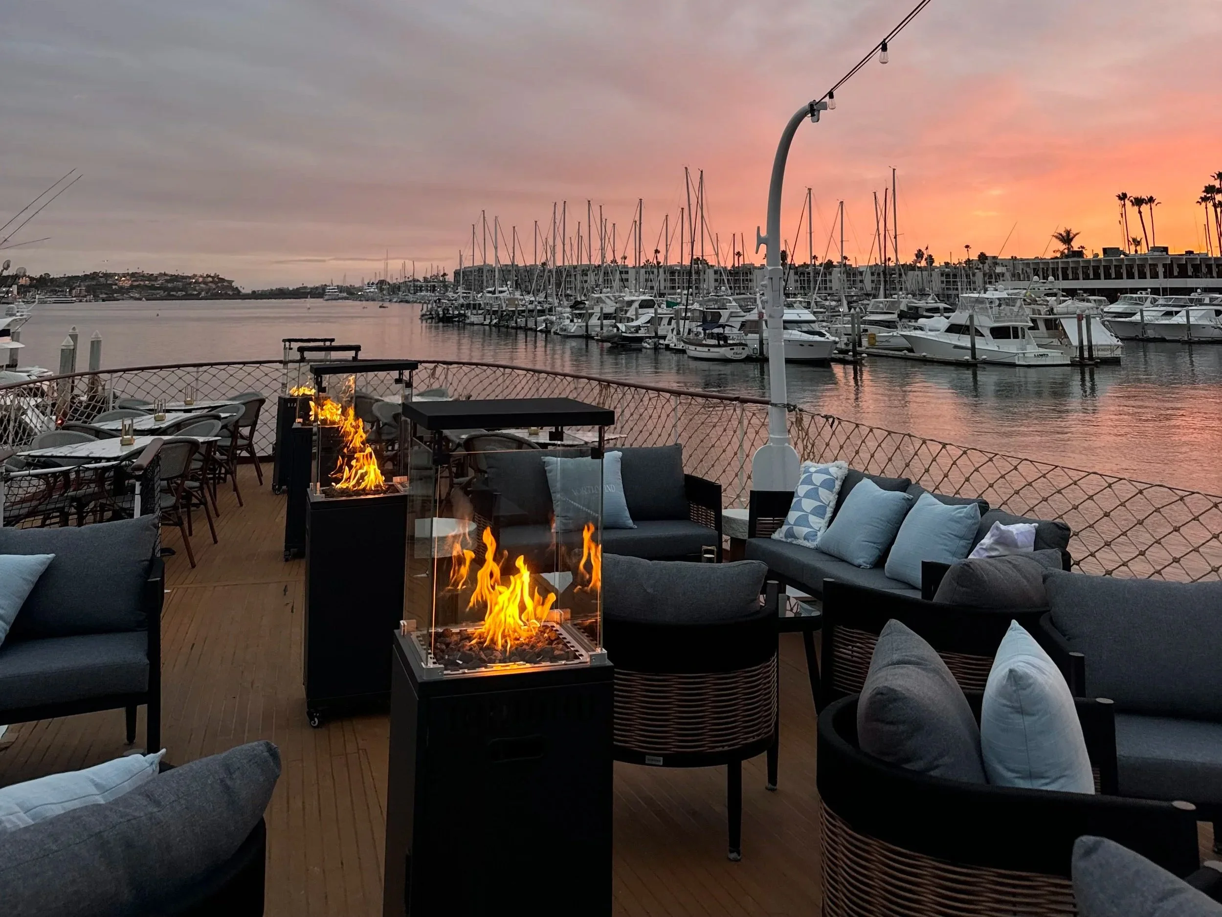 An outdoor lounge area on a boat, with white sofas and marble-top tables, overlooking a marina filled with yachts, under a clear blue sky.