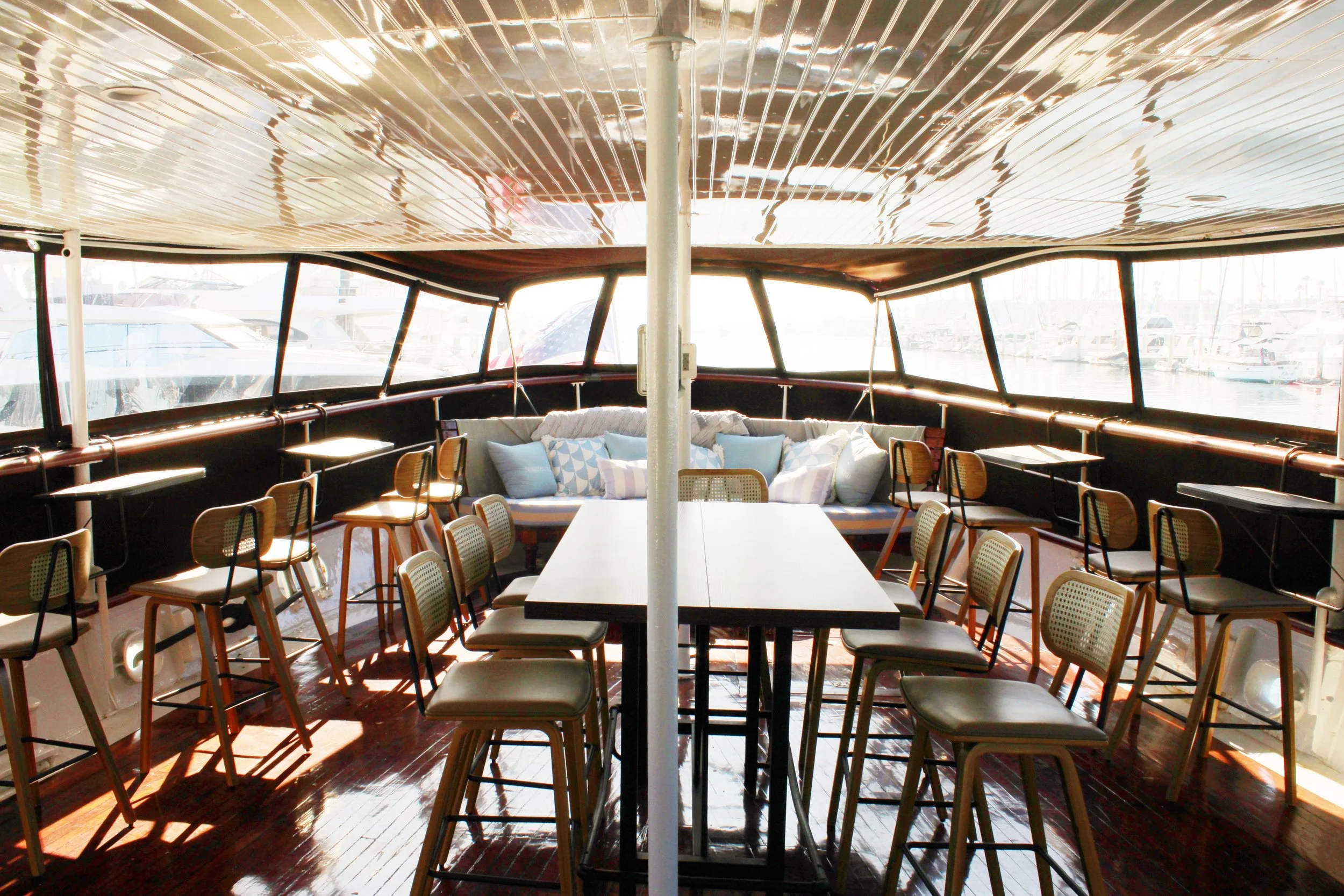 Outdoor seating area on a boat with a view of the marina and boats, American flag in the background, wooden deck, chairs, and tables.