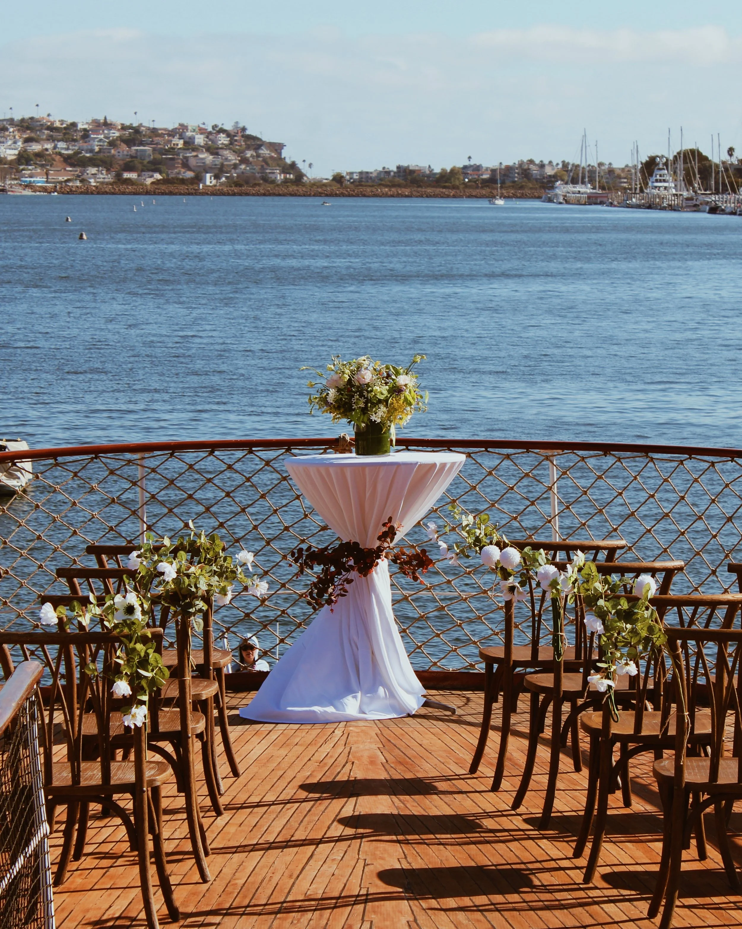 A waterfront wedding setup with a tall floral centerpiece on a high table, surrounded by chairs decorated with flowers, overlooking a body of water and a hilly cityscape in the background.