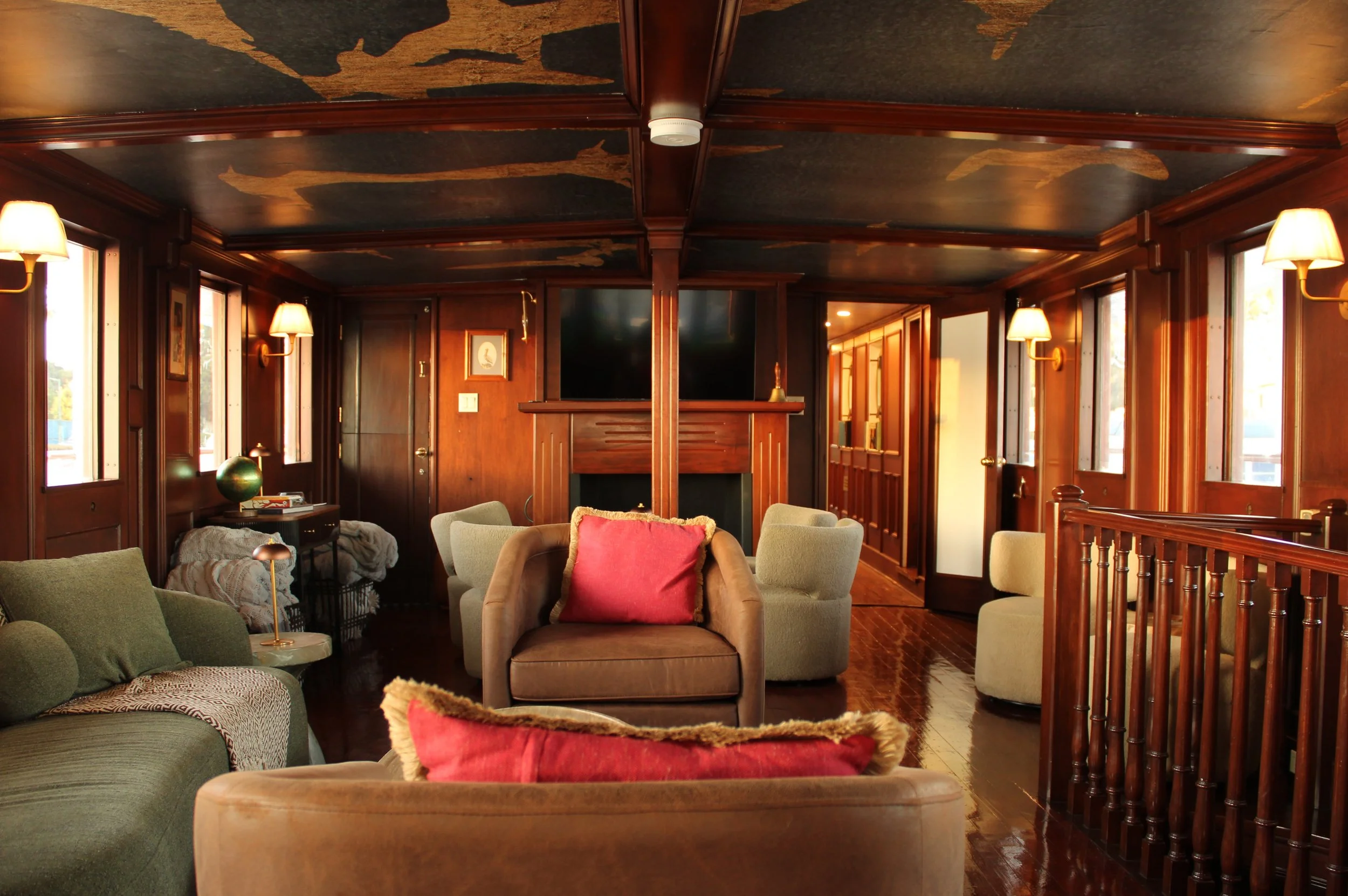 Interior of a wooden-paneled lounge room with a mural of fish on the ceiling, multiple windows, a fireplace, and a sitting area with sofas, armchairs, and a small table.