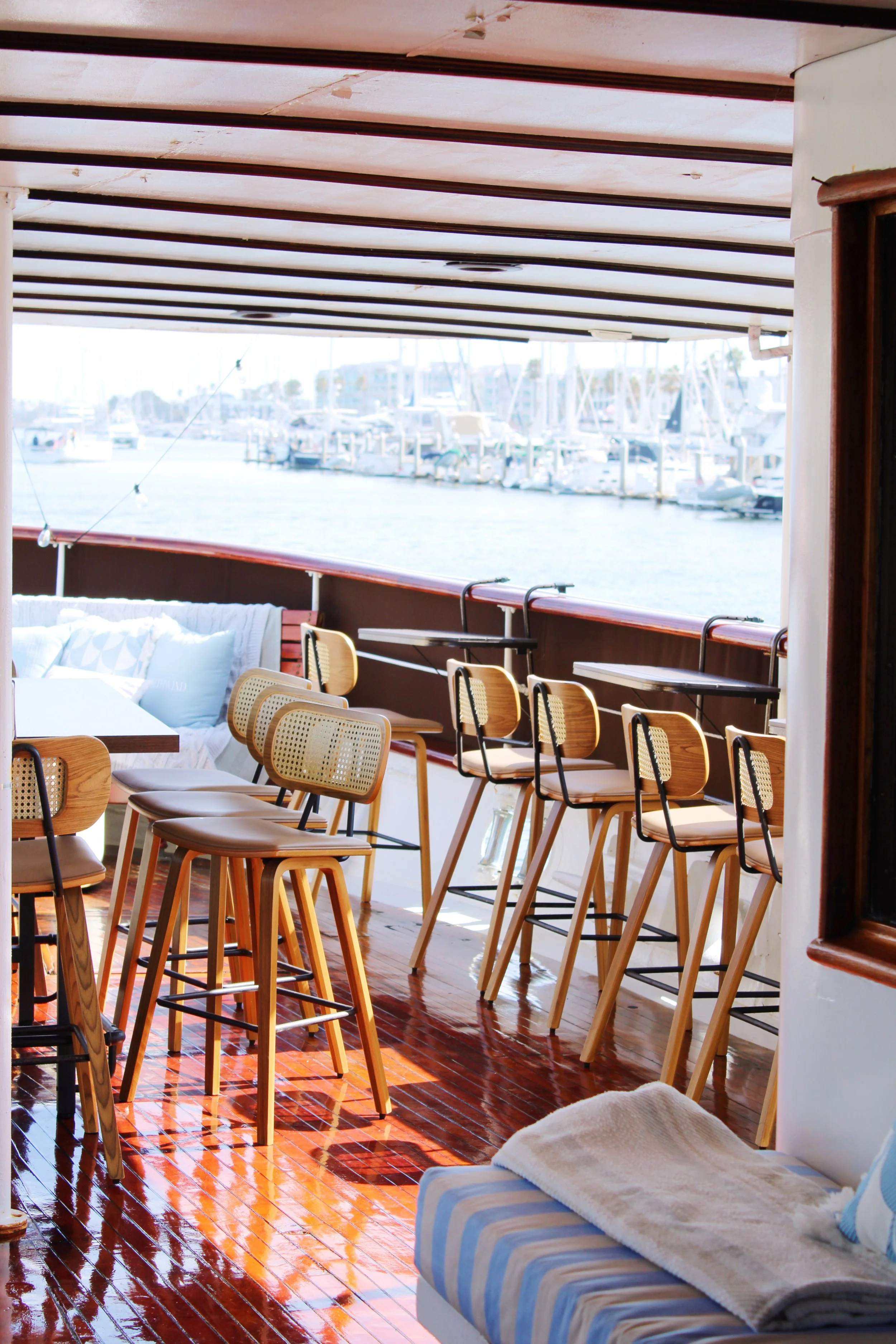 Aboard a boat, a cozy seating area with wooden chairs and a table overlooking a marina with docked boats.