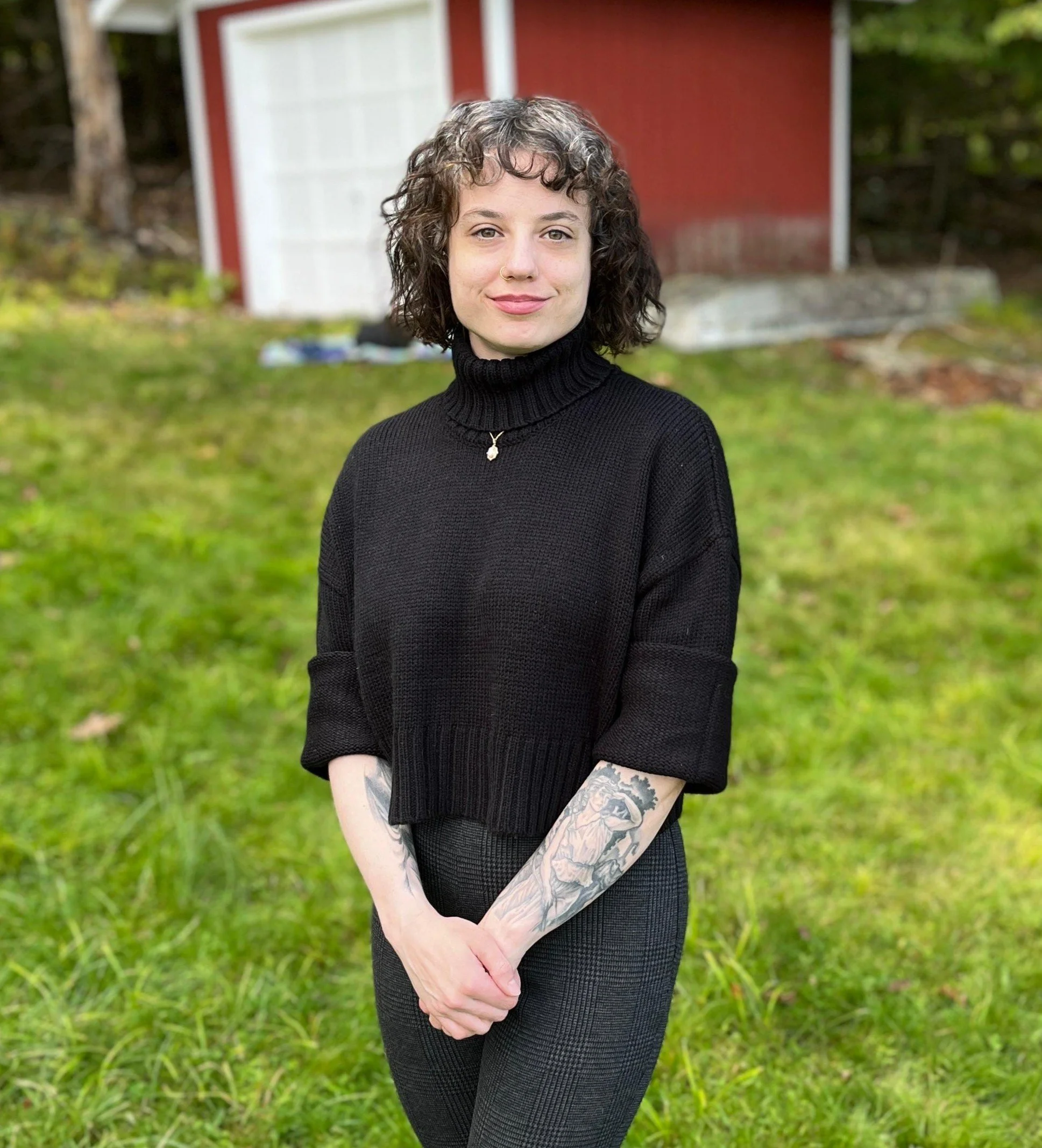 A young woman with curly short hair, wearing a black turtleneck sweater and patterned pants, standing outdoors on grass with a red barn and trees in the background.