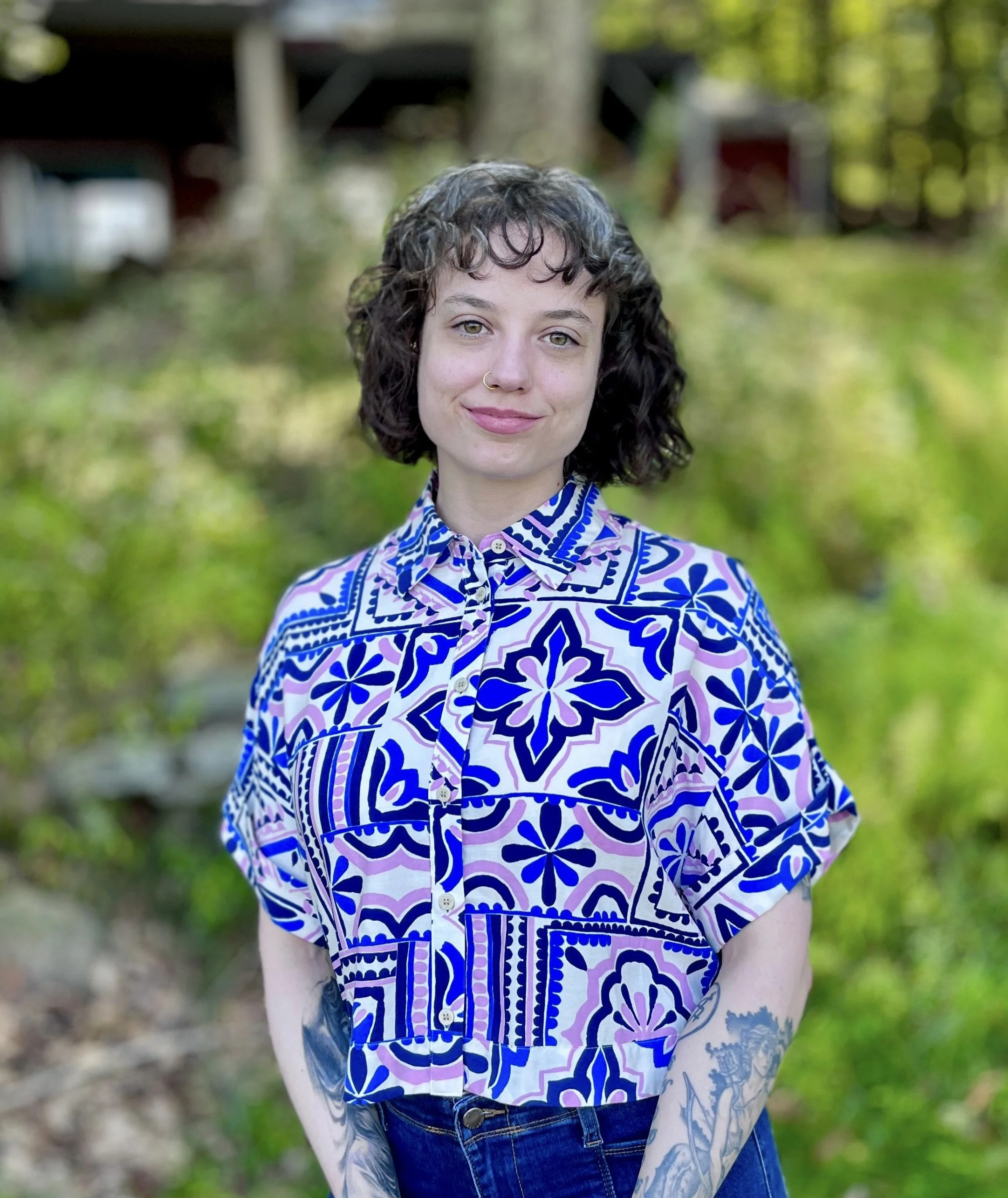 A woman who is an AASECT Certified Sex Therapist with curly dark hair and light skin standing outdoors in front of a green, leafy background. She works in person and virtually in Brooklyn, NY MA & CO