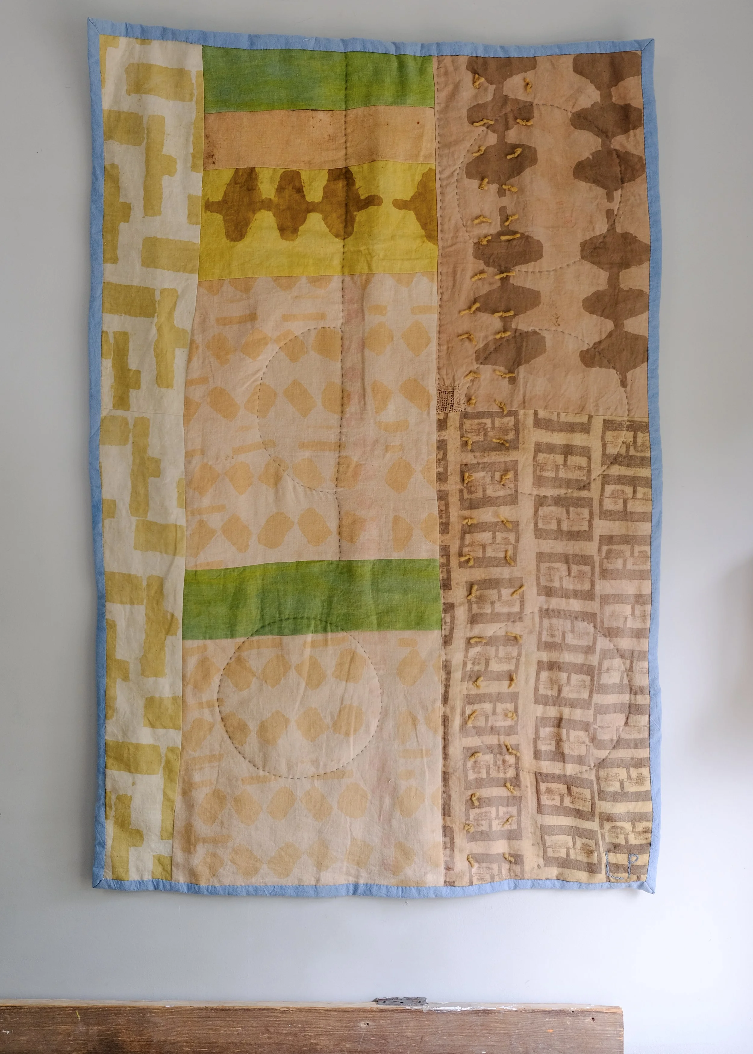 A textile wall hanging with multiple geometric and abstract patterns, bordered with light blue fabric, mounted on a white wall.