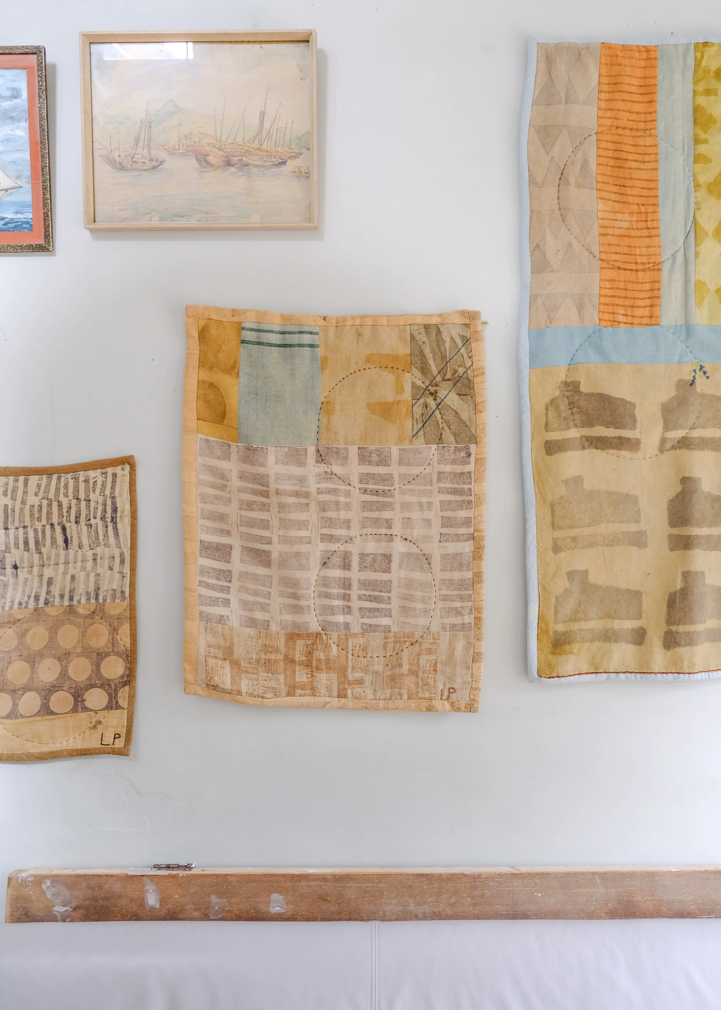 Three fabric artworks hanging on a white wall with watercolor paintings above. The artworks feature geometric shapes and patterns in earth tones, with one including a fishing reel design. A wooden support is at the bottom.