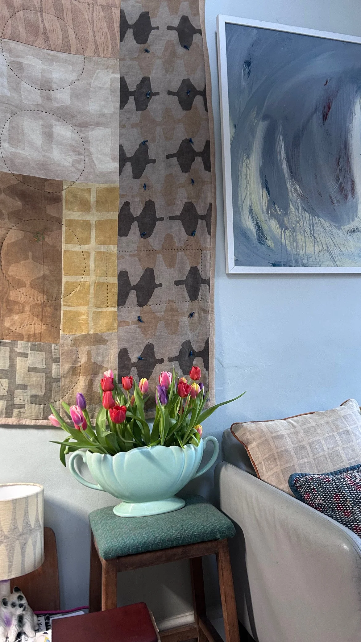 A cozy living room corner with a quilted wall hanging, a large abstract painting, a vase of pink and purple tulips, a green upholstered stool, a beige couch with patterned pillows, and a small lamp.