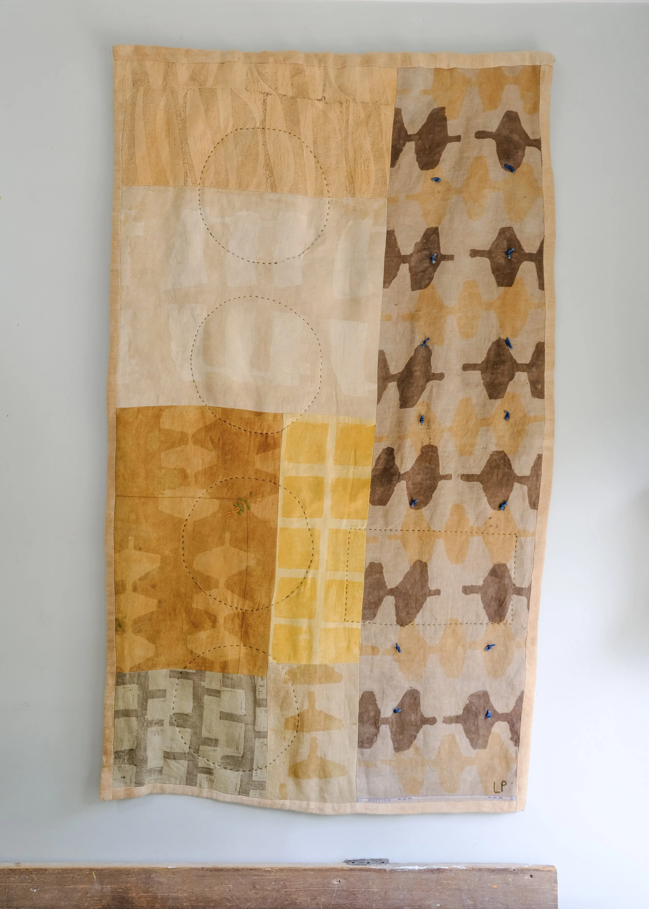 A textile wall hanging with various patchwork designs and patterns in neutral shades of beige, brown, and yellow, with some blue pins holding it to the wall.