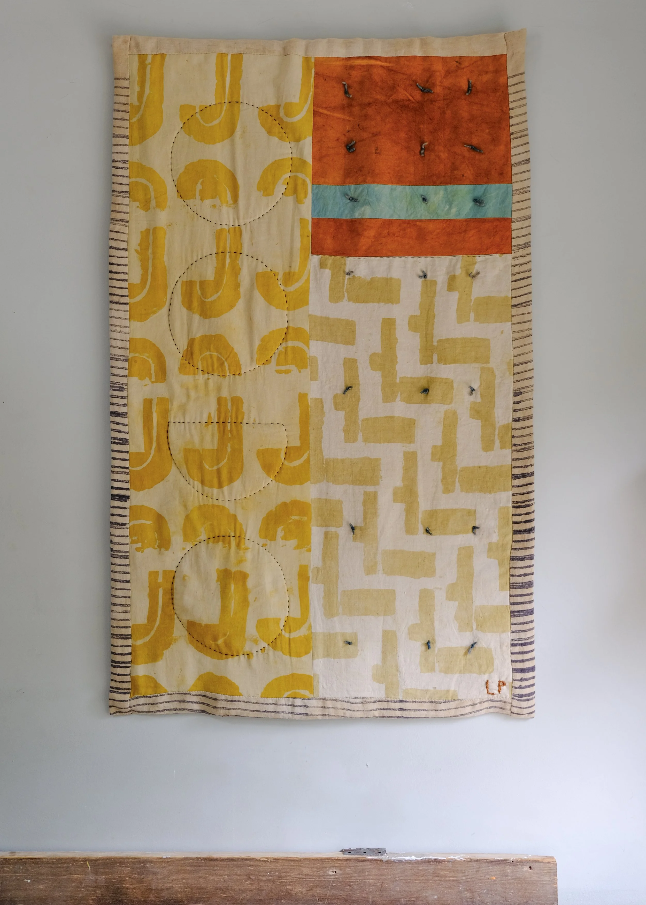 A textile wall hanging with abstract shapes and patterns in yellow, orange, and blue, with visible hooks securing it to the wall.