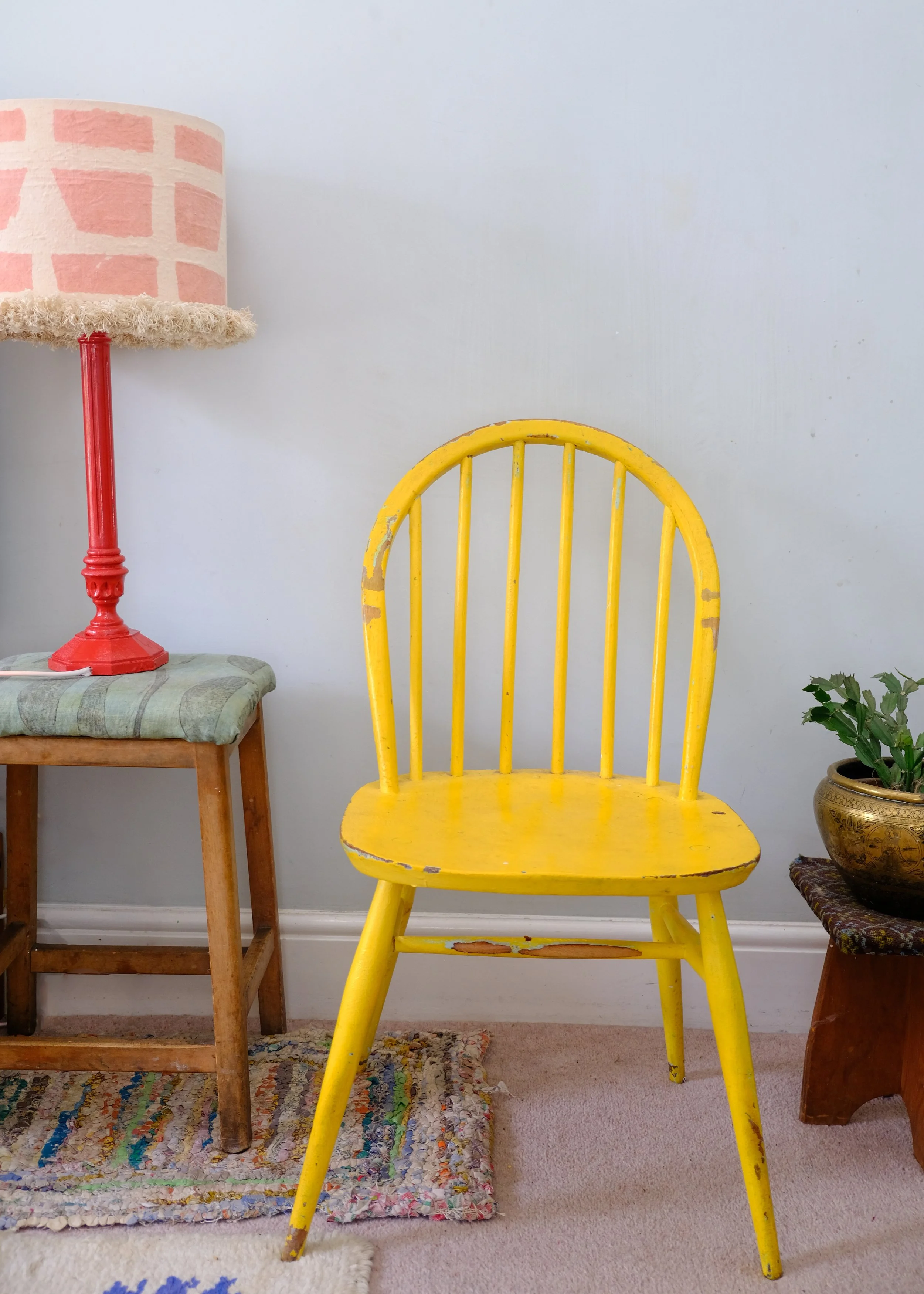 A yellow wooden chair with a distressed finish, located in a room with a colorful braided area rug, a small table with a potted plant, a wooden stool with a cushion, and a pink and white table lamp on a side table.