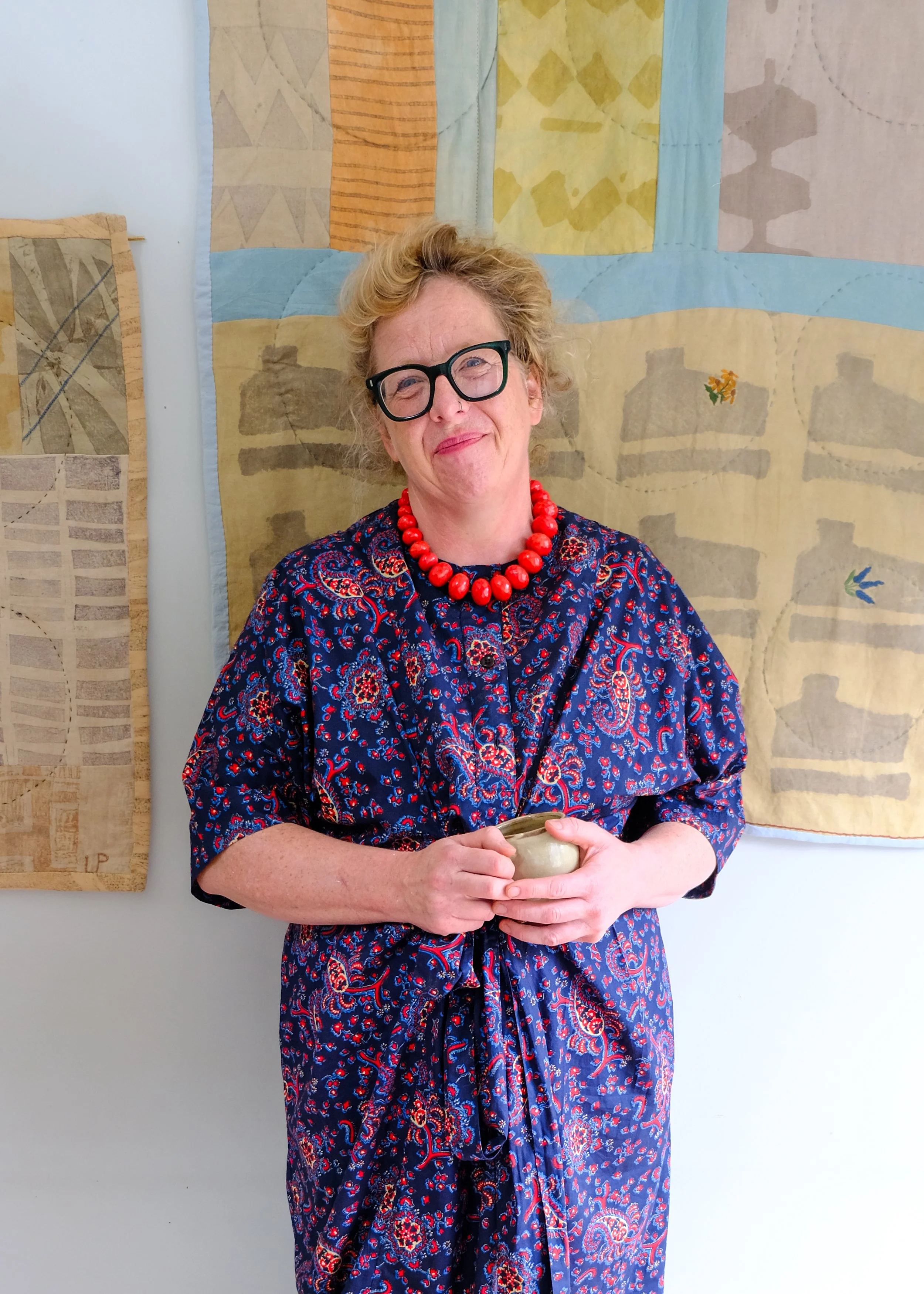 A woman with curly hair, wearing glasses, a red necklace, and a navy blue dress with red and white patterns, holding a small bowl, standing in front of a quilt with geometric patches and various muted colors.