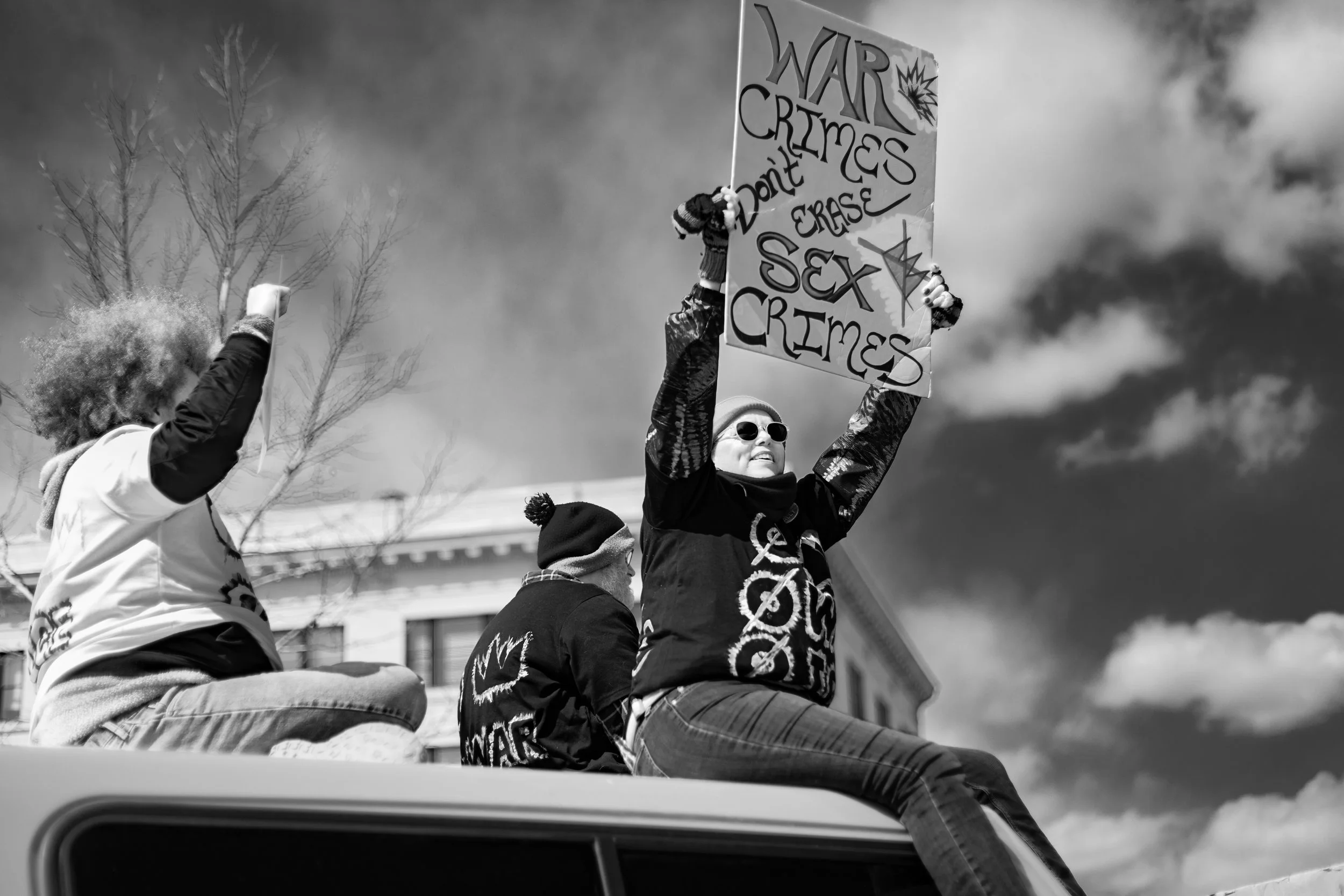 Group of protesters on top of a vehicle holding signs that read "War Crimes Don't Erase Sex Crimes" during a rally or demonstration, with a cloudy sky in the background.