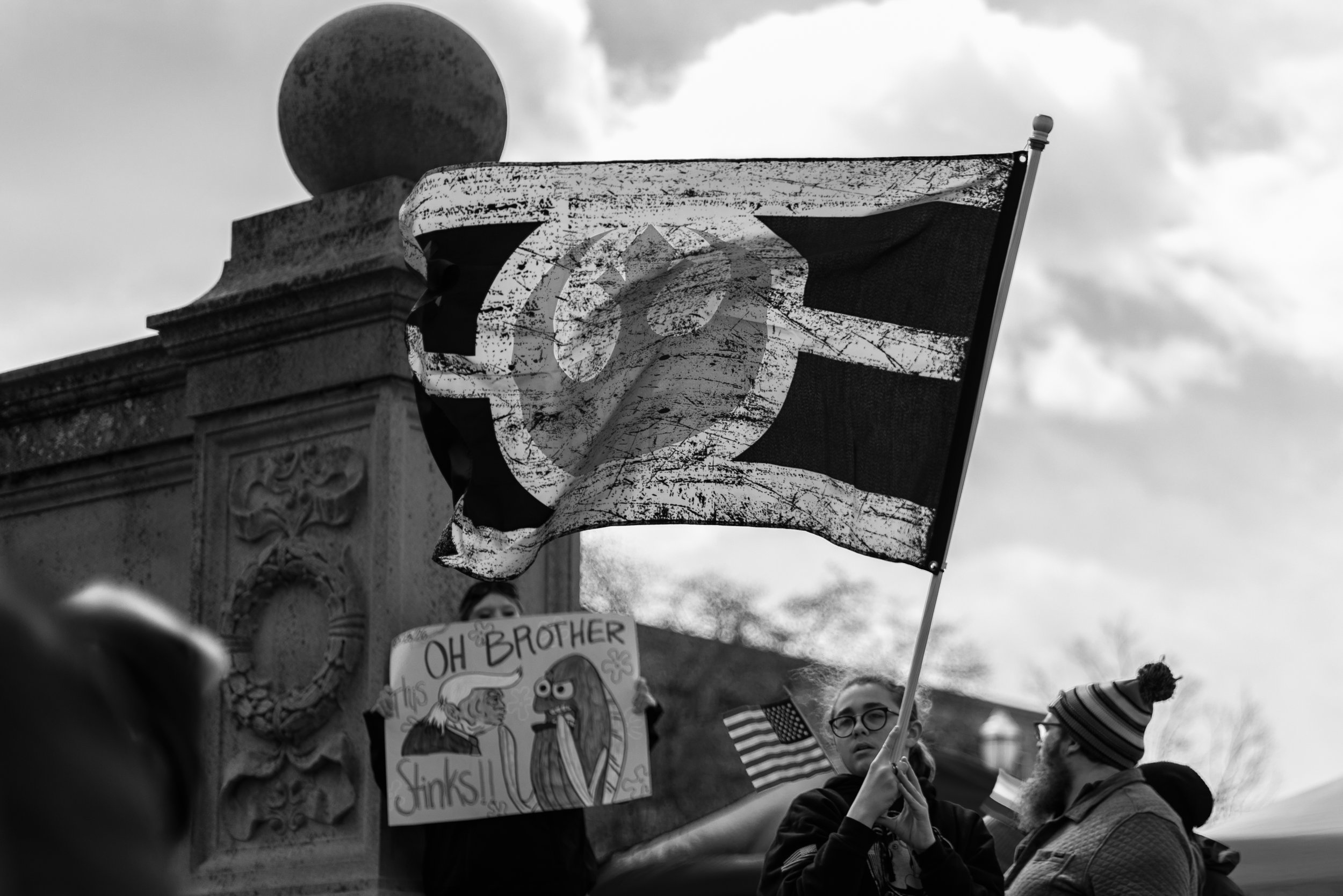 Black and white photo of children holding signs and flags at a protest or rally, with part of an ornate stone monument visible in the background. One sign reads "OH BROTHER" and features cartoon characters, including Sesame Street's Elmo and a lion.
