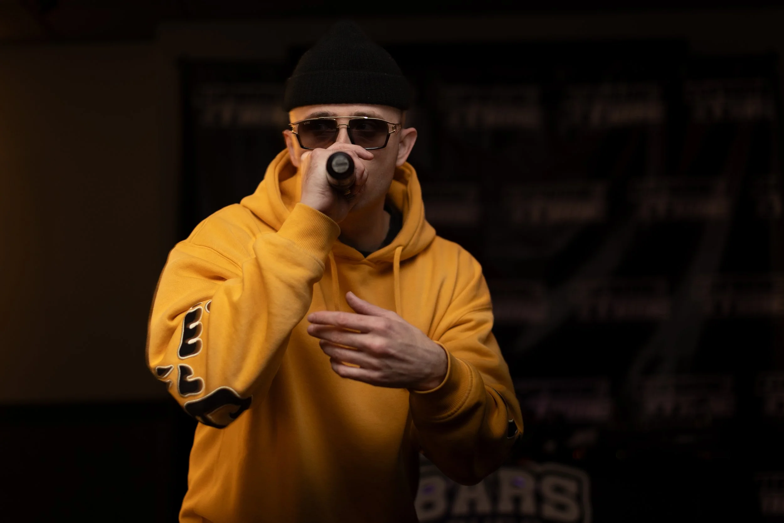 A young man wearing a yellow hoodie, black beanie, and sunglasses holding a microphone and singing or speaking on stage.
