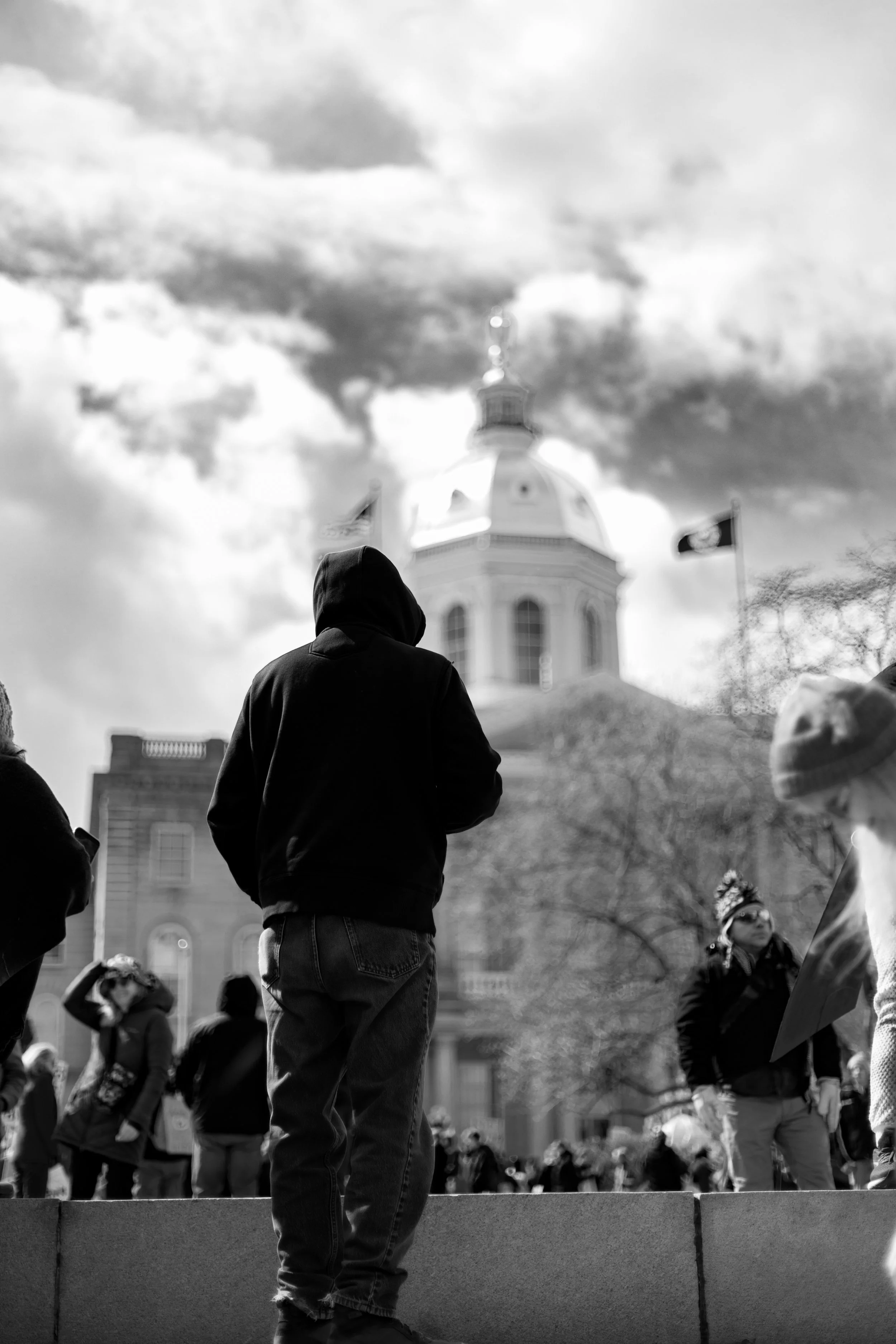 A black and white photo of a crowd of people gathered outdoors in front of a government building with a dome and a flag flying nearby. The focus is on the back of a person wearing a hoodie and jeans, standing on a raised platform.