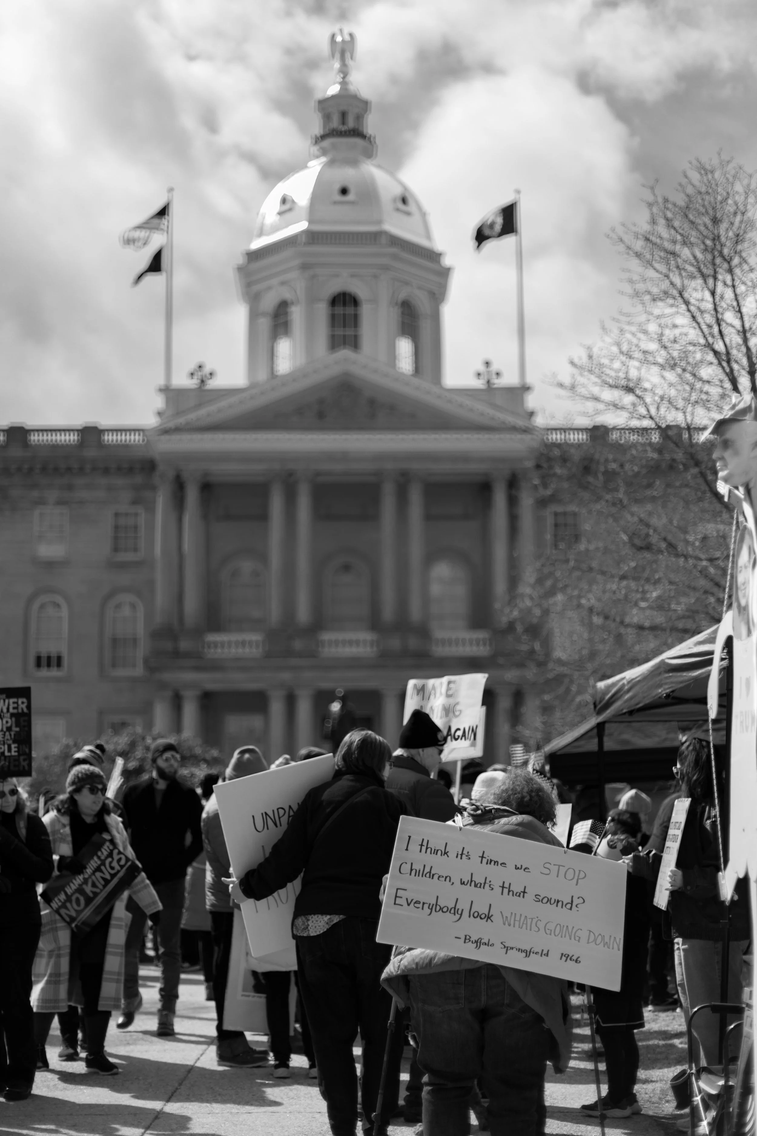 A group of people participating in a protest or rally outside a government building with a domed roof and flags flying. Many are holding signs with messages.