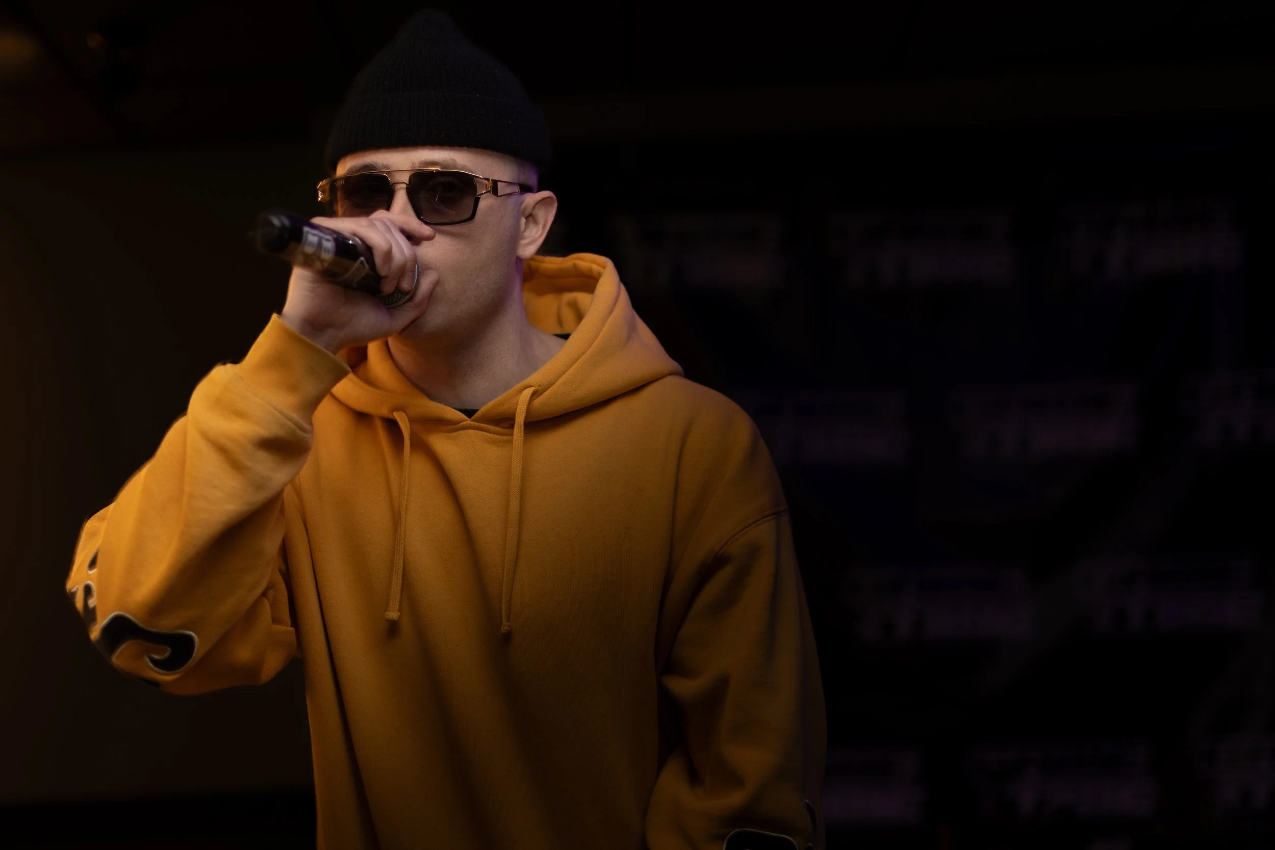A person wearing a yellow hoodie, black beanie, and sunglasses is holding a microphone in their hand.
