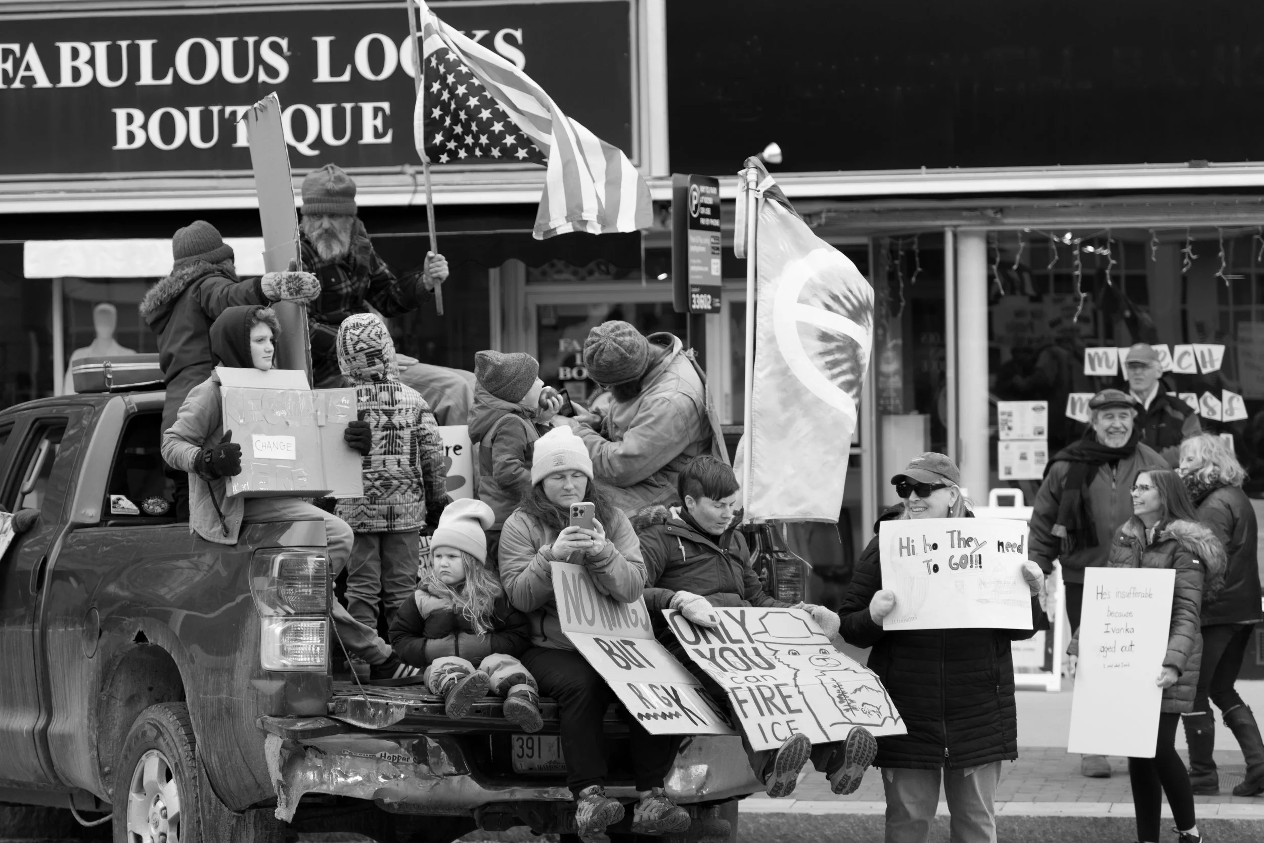 A group of people gathered in a protest, some holding signs and flags. Several individuals are sitting on the back of a pickup truck, with one man holding an American flag and a woman holding a sign. The background shows storefronts and additional pr