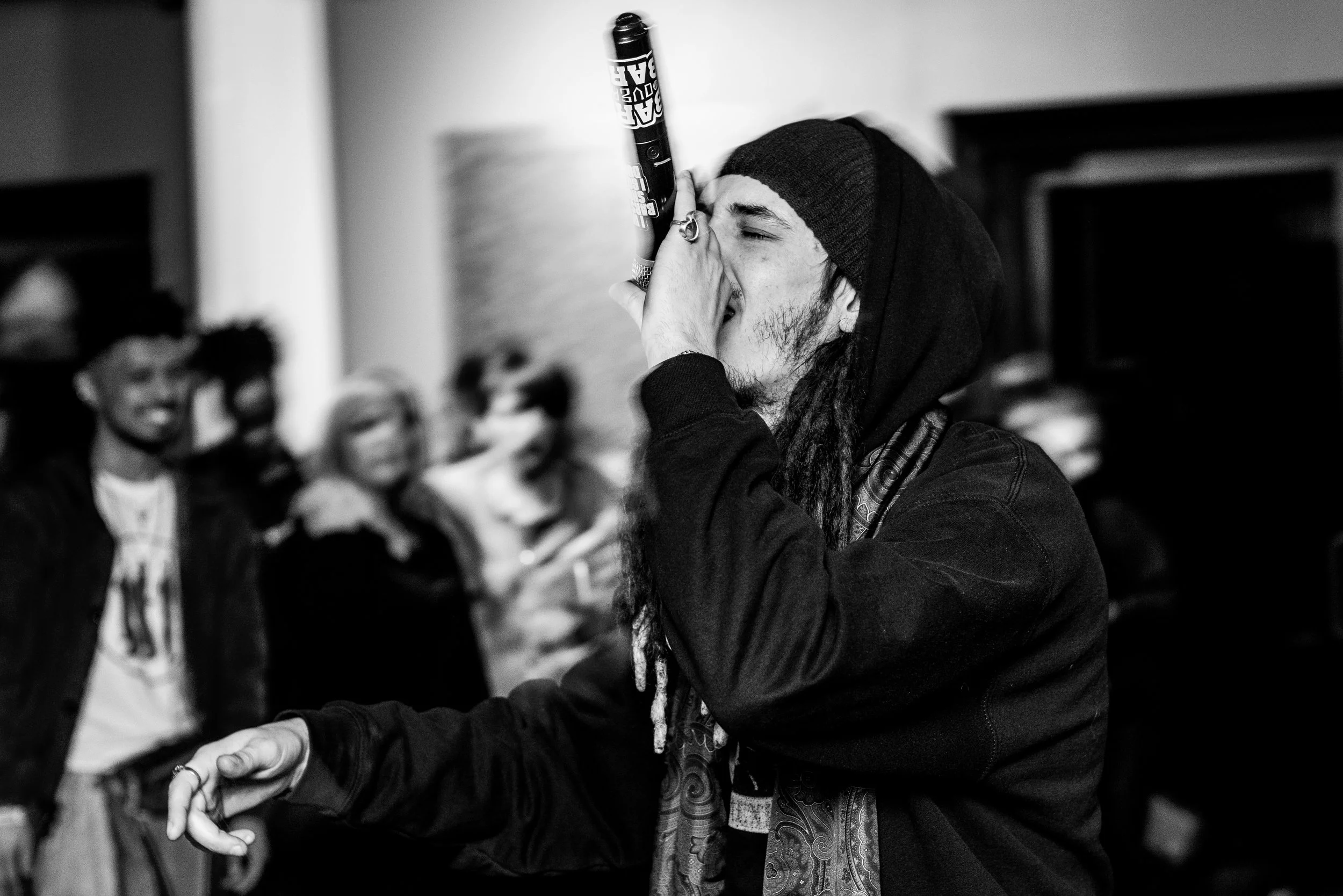 A person with long hair, wearing a knit cap and a jacket, holding a marker and smoking a joint, surrounded by a group of people in an indoor setting.