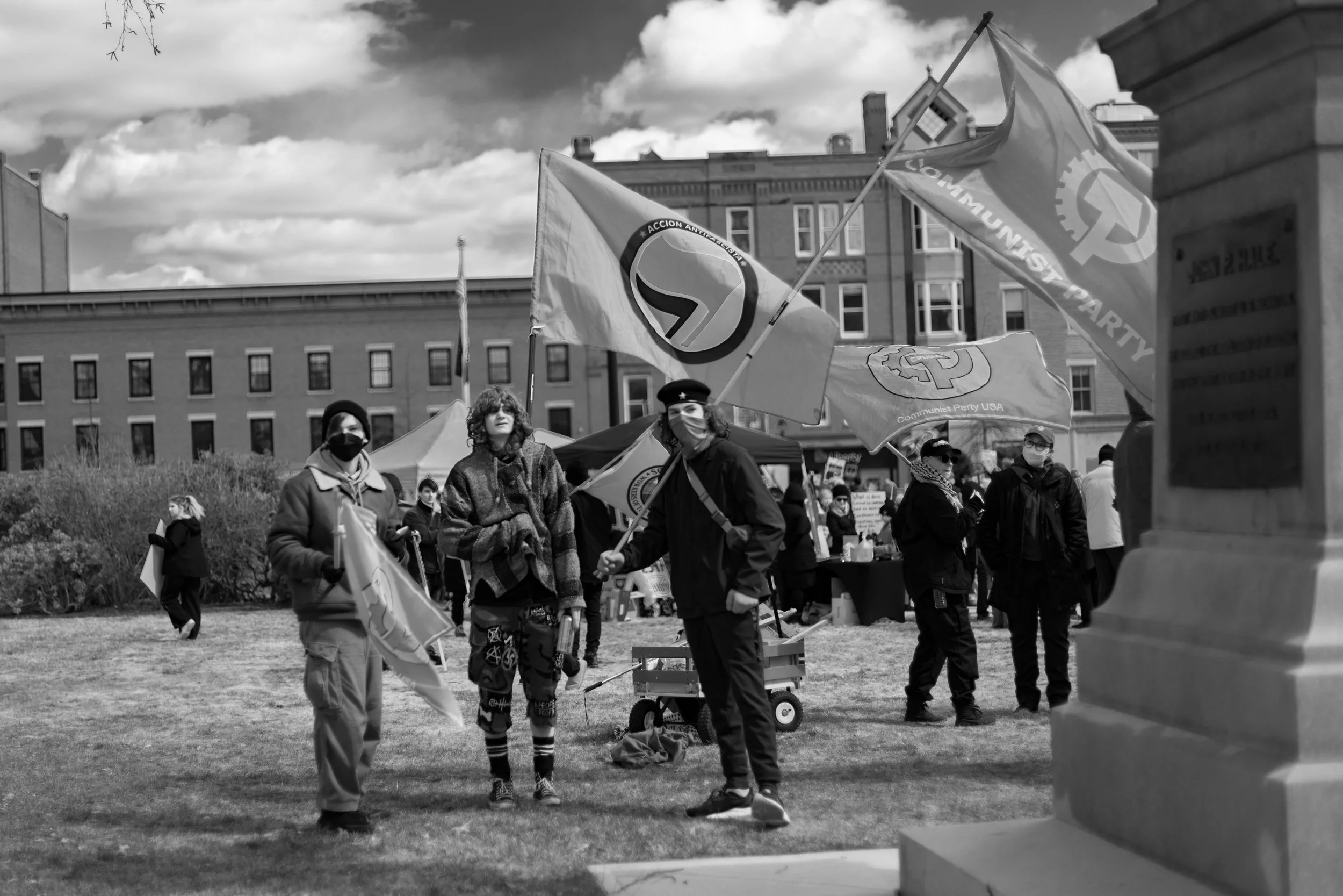 People participating in a protest or rally holding flags, with some wearing masks and standing on a grassy area in front of buildings, a tent, and clouds in the sky.