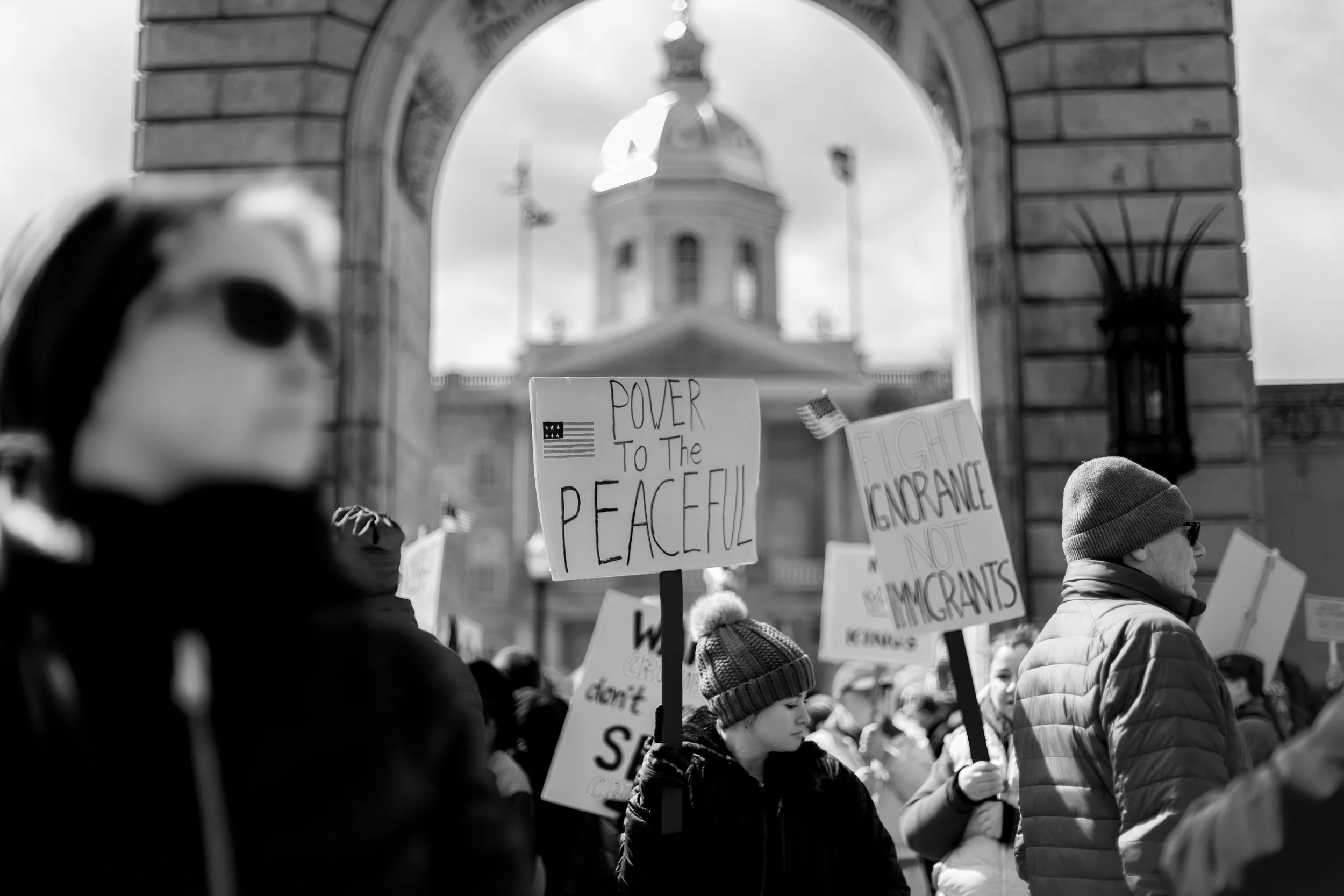 People participating in a protest or demonstration, holding signs with messages such as "Power to the Peaceful" and "Fight Ignorance, Not Immigrants," in front of a government building or monument.