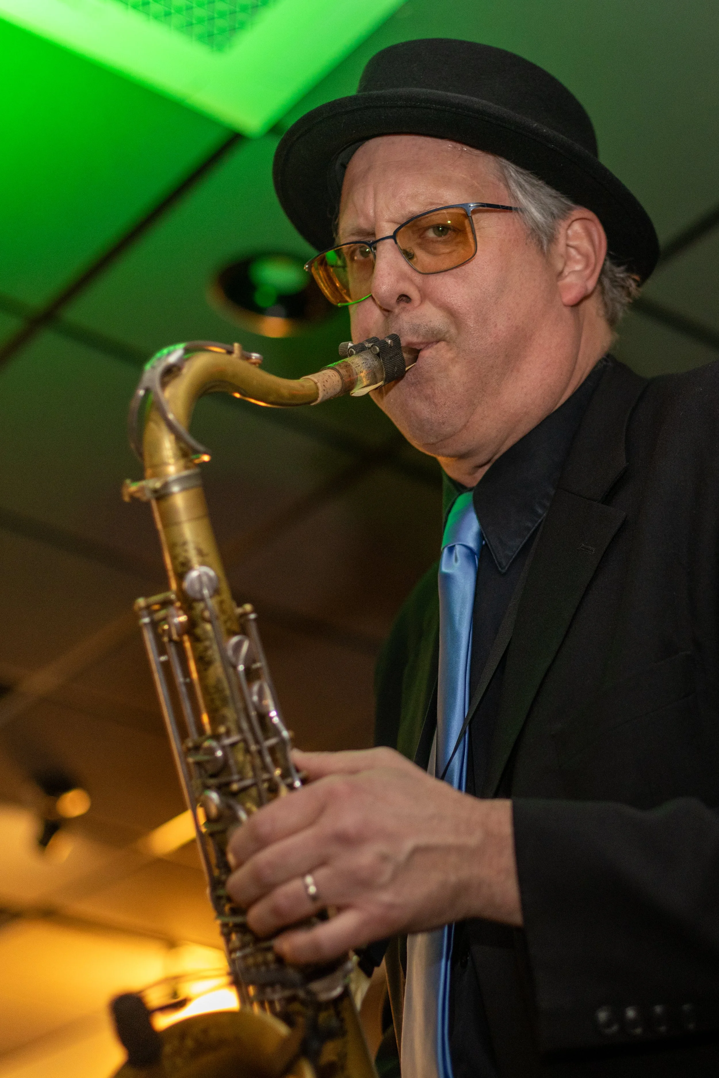 A man wearing a black hat, glasses, a black suit, and a blue tie playing a saxophone in a room with green lighting.