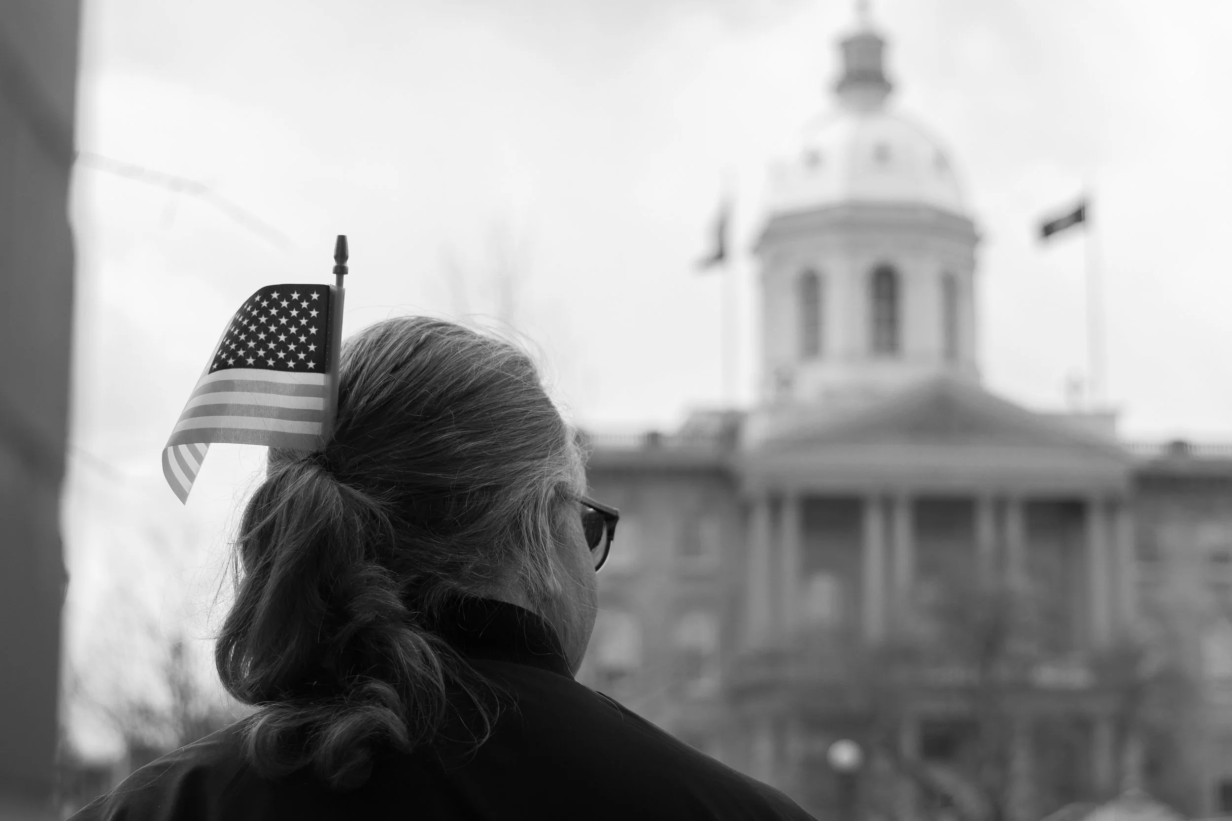 A woman with curly hair and glasses is seen from the side, with an American flag in her hair, overlooking a historic building with a dome in the background, all in black and white.
