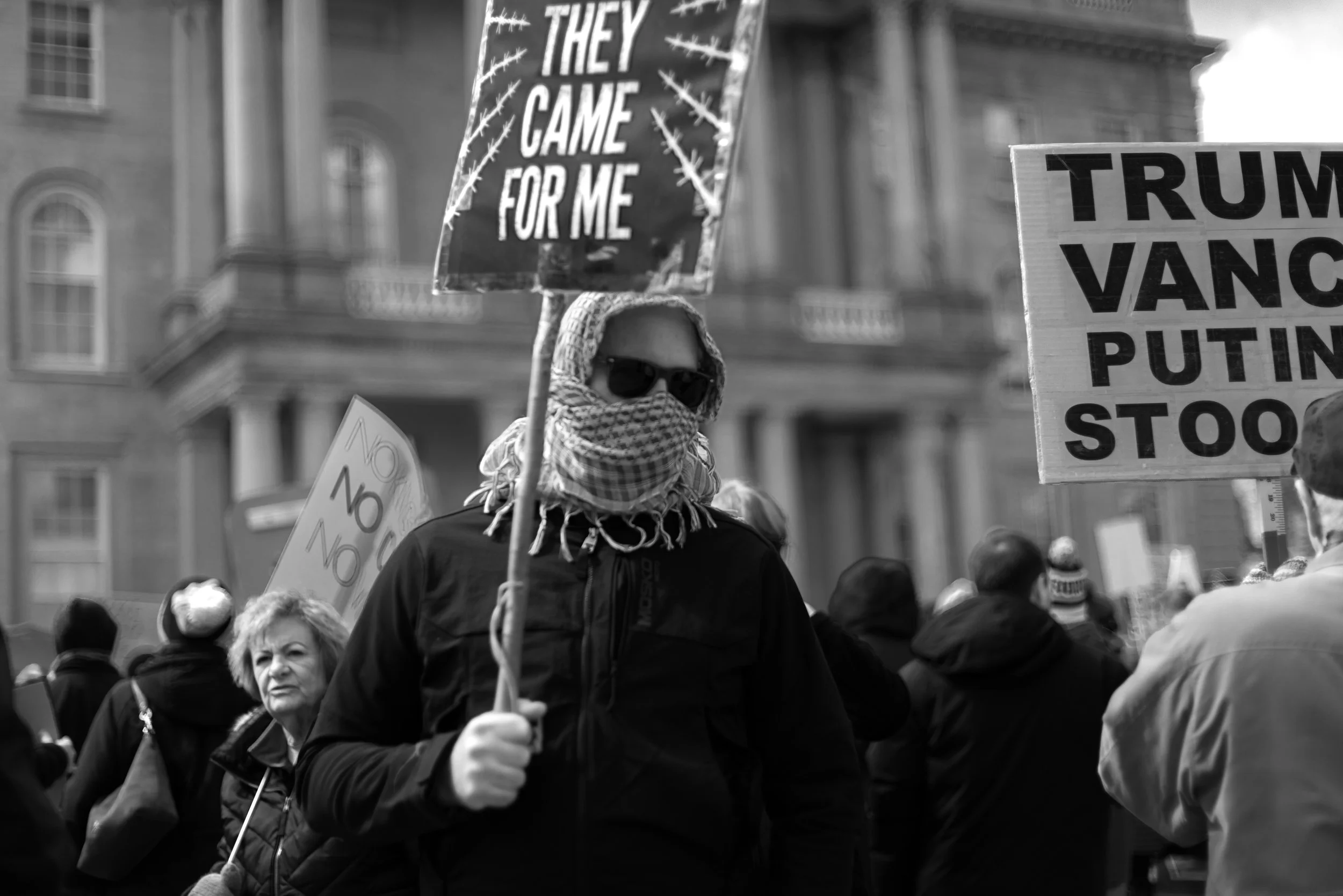 A person wearing sunglasses and a scarf covering their face, holding a sign that reads 'They Came for Me' during a protest or rally in a city setting with multiple signs and a crowd of people.