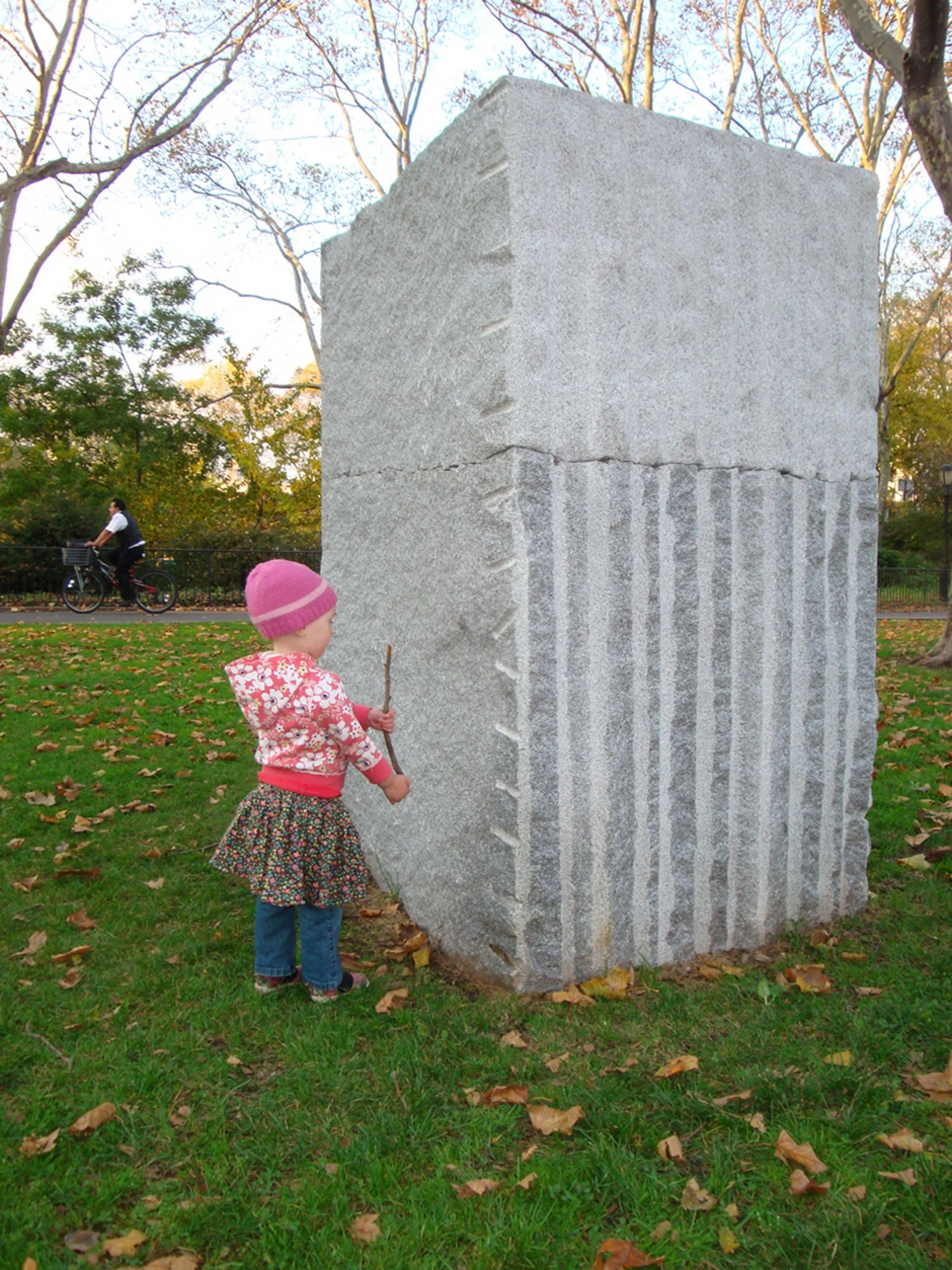 Rear view of a large granite sculpture in a park, with a child standing beside it holding a stick.