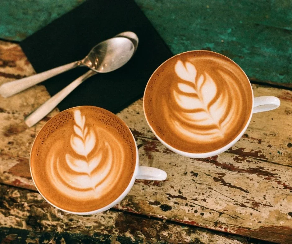 Two cups of coffee with latte art on a rustic wooden table, along with a spoon and napkin.