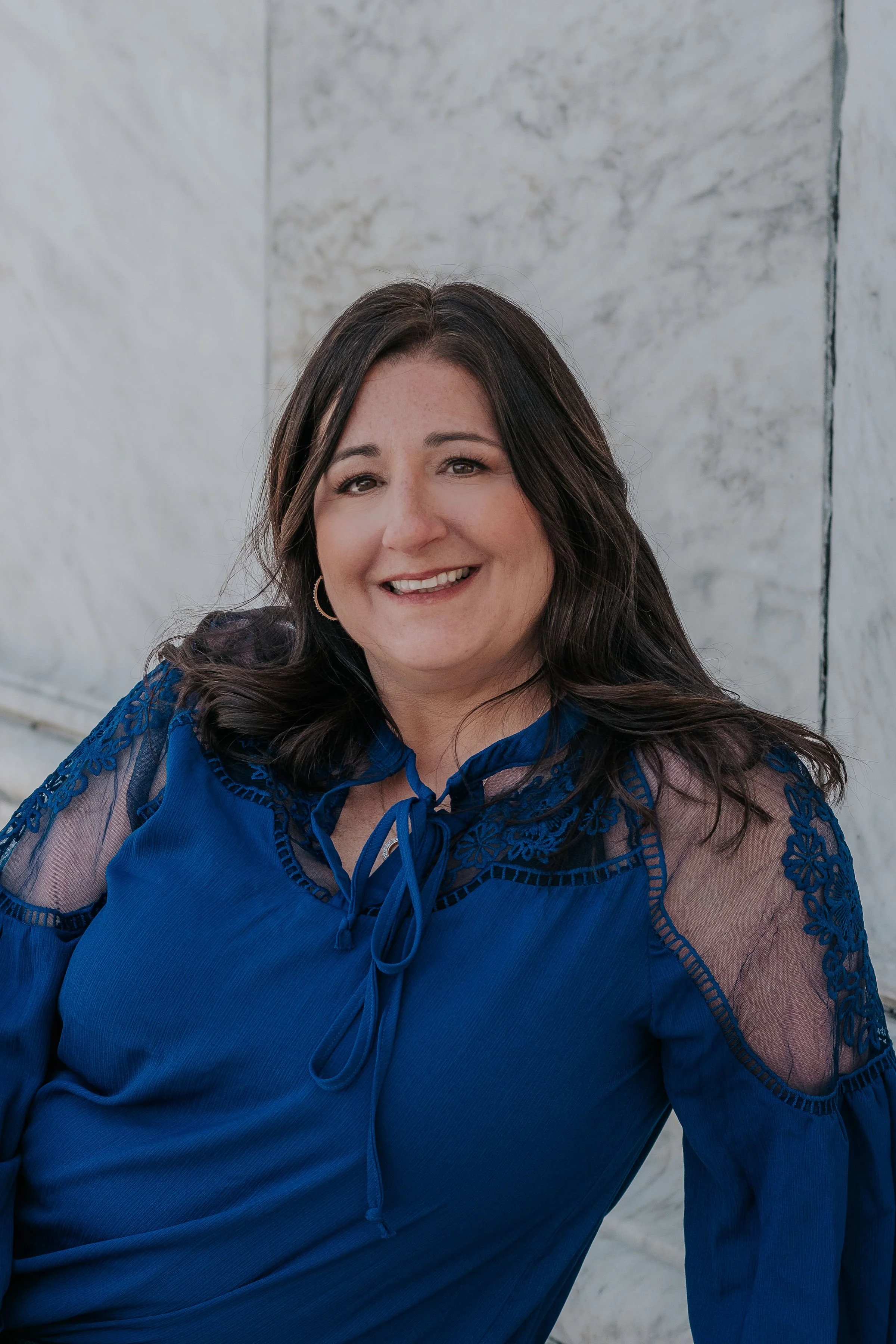 Smiling woman with dark brown hair in a blue dress with lace details, standing in front of a marble wall.