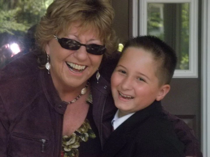An elderly woman wearing dark sunglasses and jewelry is hugging a young boy with short dark hair. Both are smiling happily and appear to be outside near a house.