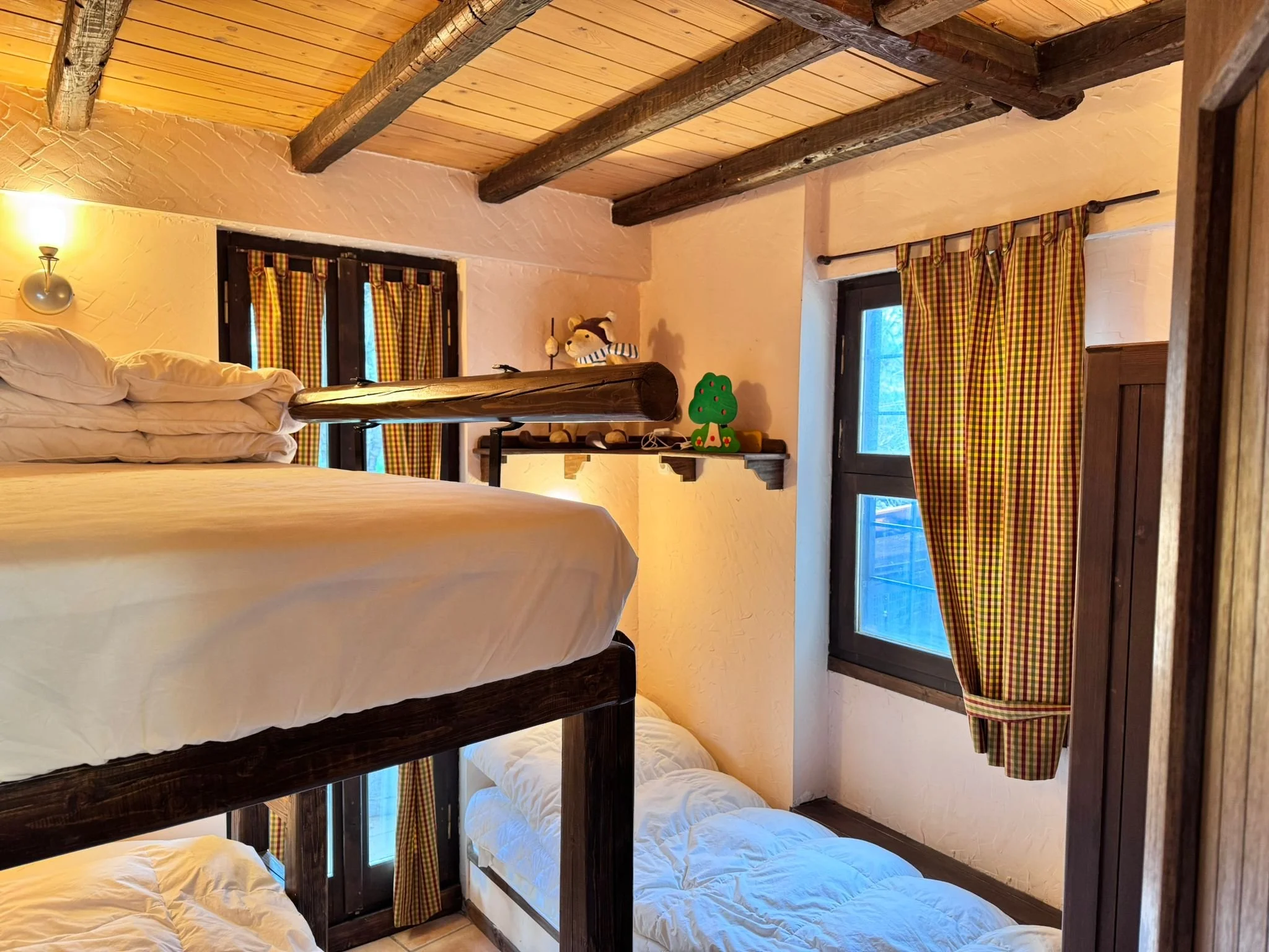 A cozy room with bunk beds, featuring pillows and white bedding. The room has textured walls, wooden ceiling beams, and windows with checkered red, yellow, and green curtains. There is a shelf with a small plush toy and a decorative green tree. Warm 