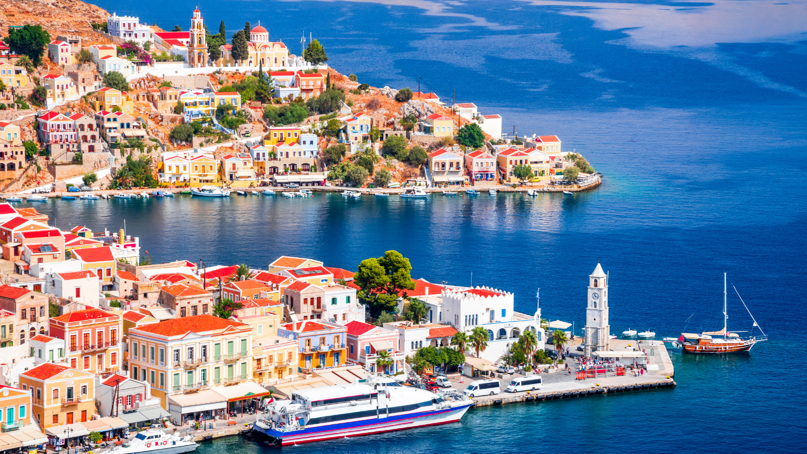 Colorful buildings with red roofs along a hillside and waterfront in a coastal town, featuring a church, clock tower, boats, and a large ferry docked by a pier.