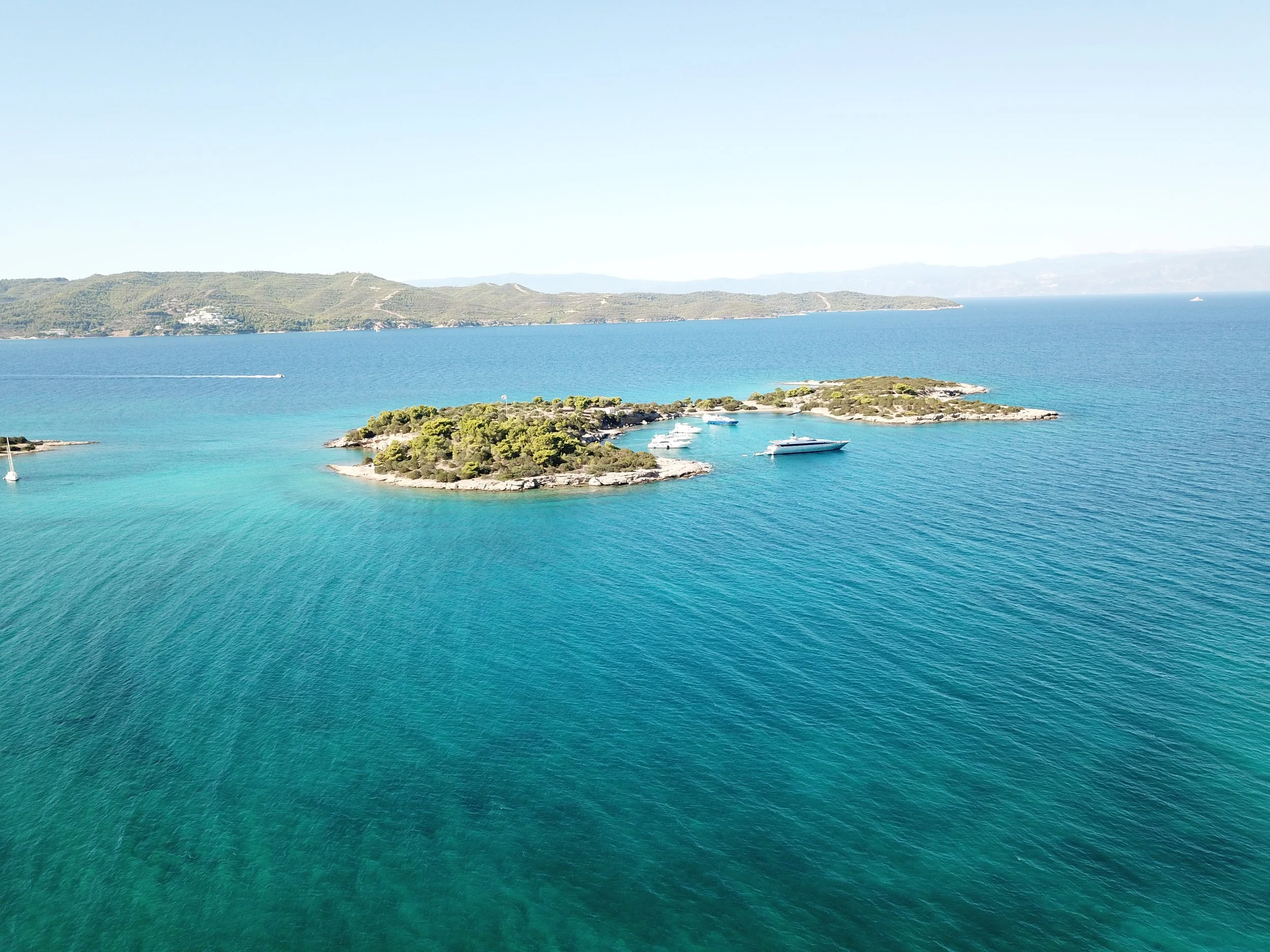 Aerial view of a small island with green trees and rocky shores, surrounded by blue ocean water, with boats docked near the island and distant hills on the horizon under a clear sky.