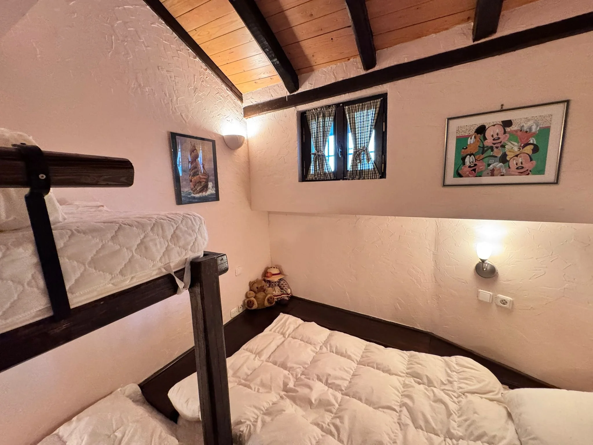 A cozy bedroom with a bunk bed and a double bed, decorated with Disney-themed artwork, including Mickey Mouse and other characters, on textured beige walls with a window and curtains, teddy bears on the corner, and warm lighting.