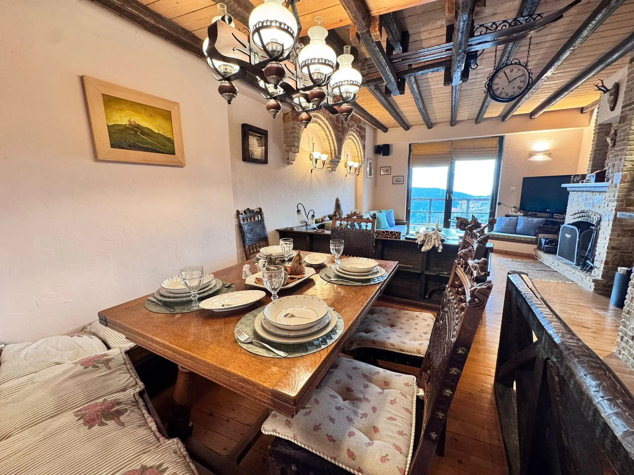A rustic dining room with a wooden dining table set with plates, bowls, glasses, and napkins, surrounded by upholstered chairs. The room features exposed wooden beams on the ceiling, brick accents, a fireplace, and a large window with a view outside.