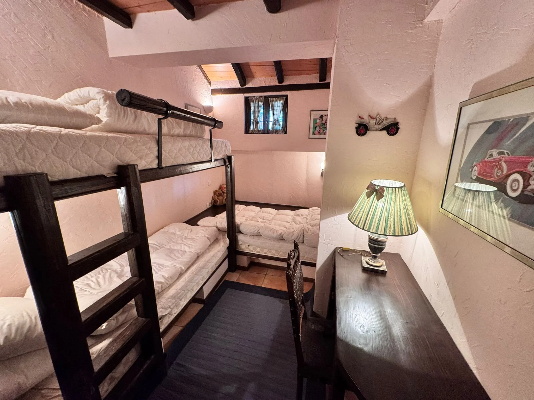 Cozy bedroom with a wooden desk, a table lamp with a green and white striped shade, a framed picture of a vintage car on the wall, and bunk beds with white bedding. There are small decorative items on the corner of the beds and a window with black la