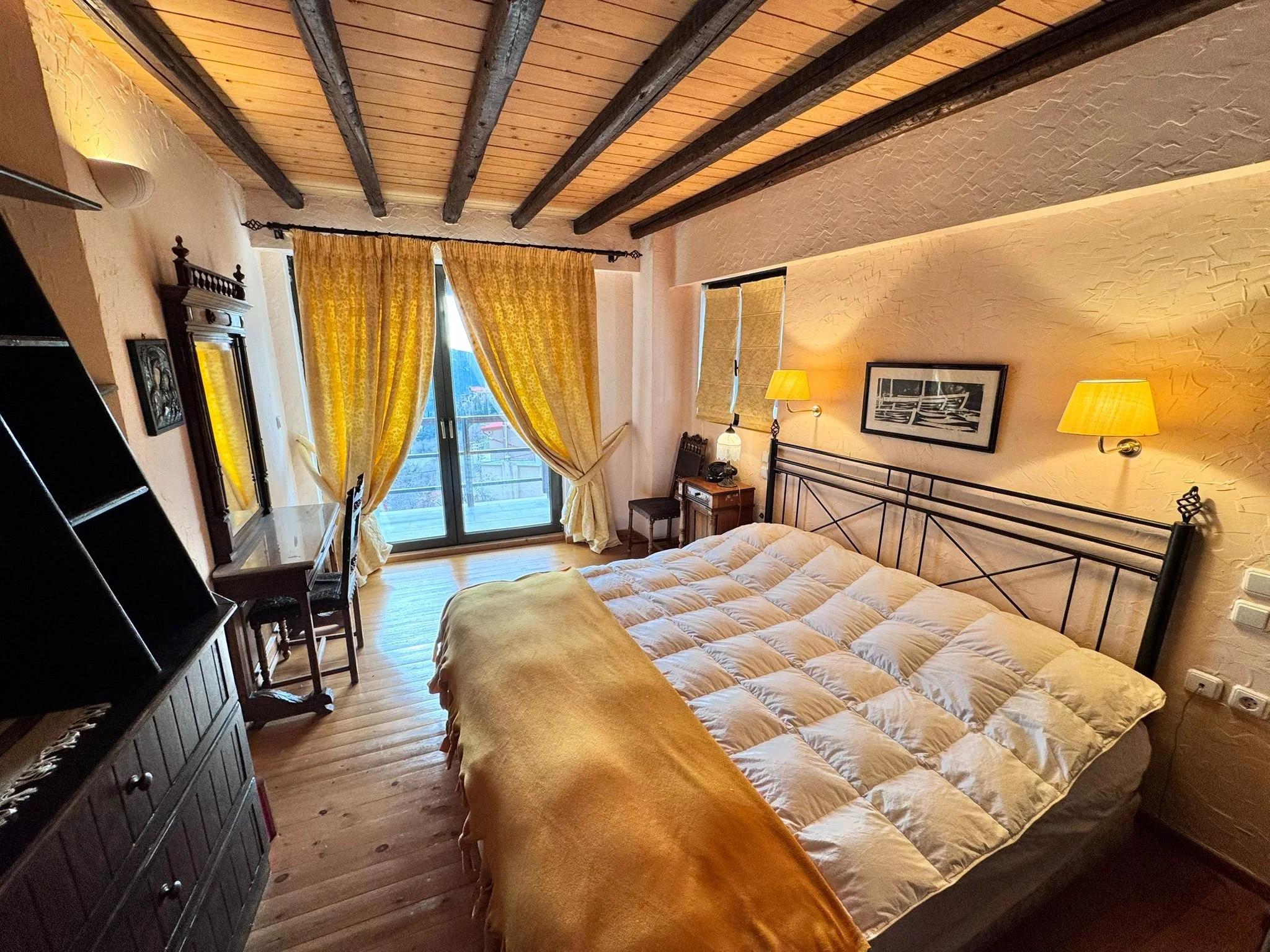 A cozy bedroom with wooden flooring, yellow curtains, and a metal bed frame. There are two yellow wall-mounted lamps and a framed picture above the bed. A small desk with a mirror and chair is near the window, which is partially open with a balcony o