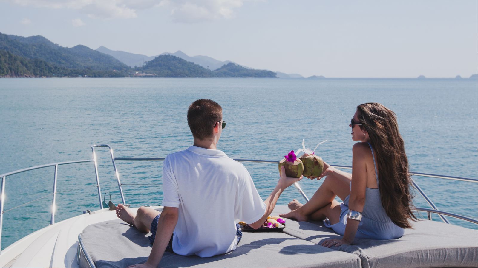 A couple relaxing on a yacht, sharing drinks from coconuts, with an ocean and islands in the background.
