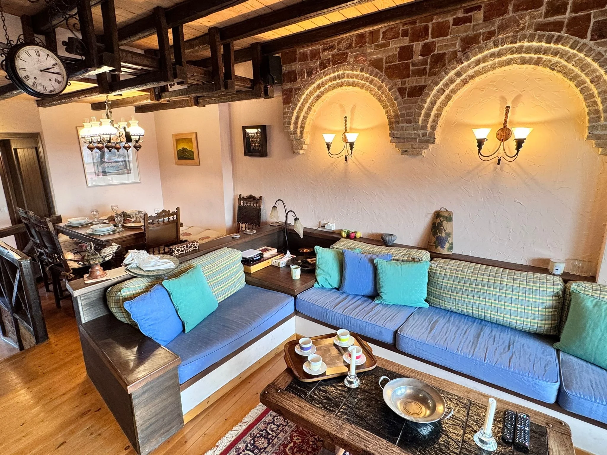 Living room with rustic brick walls, arched brick accents, wall-mounted lamps, wooden furniture, a sectional sofa with colorful cushions, a wooden coffee table with cups, a dining area with a chandelier, and decorative items.