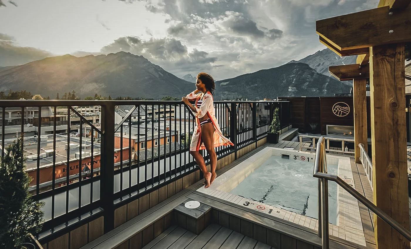 A woman in a swimsuit and a colorful cover-up stands on a balcony overlooking a mountain range and a city below, next to a hot tub.